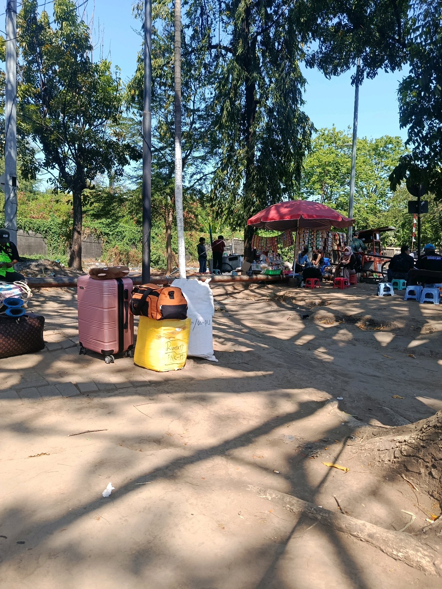 The image shows an outdoor scene in front of the Bungurasih bus station in Surabaya, Indonesia. In the foreground, there are several pieces of luggage, including a pink suitcase, a yellow plastic bag, and a brown designer-patterned bag. Behind the luggage, a red umbrella shades a food stall where several people are seated on small plastic stools, purchasing and consuming food and drinks. A few individuals are standing or walking around the stall. The environment is characterized by dirt ground, trees, and utility poles. The overall impression is a bustling, everyday scene at a transportation hub.