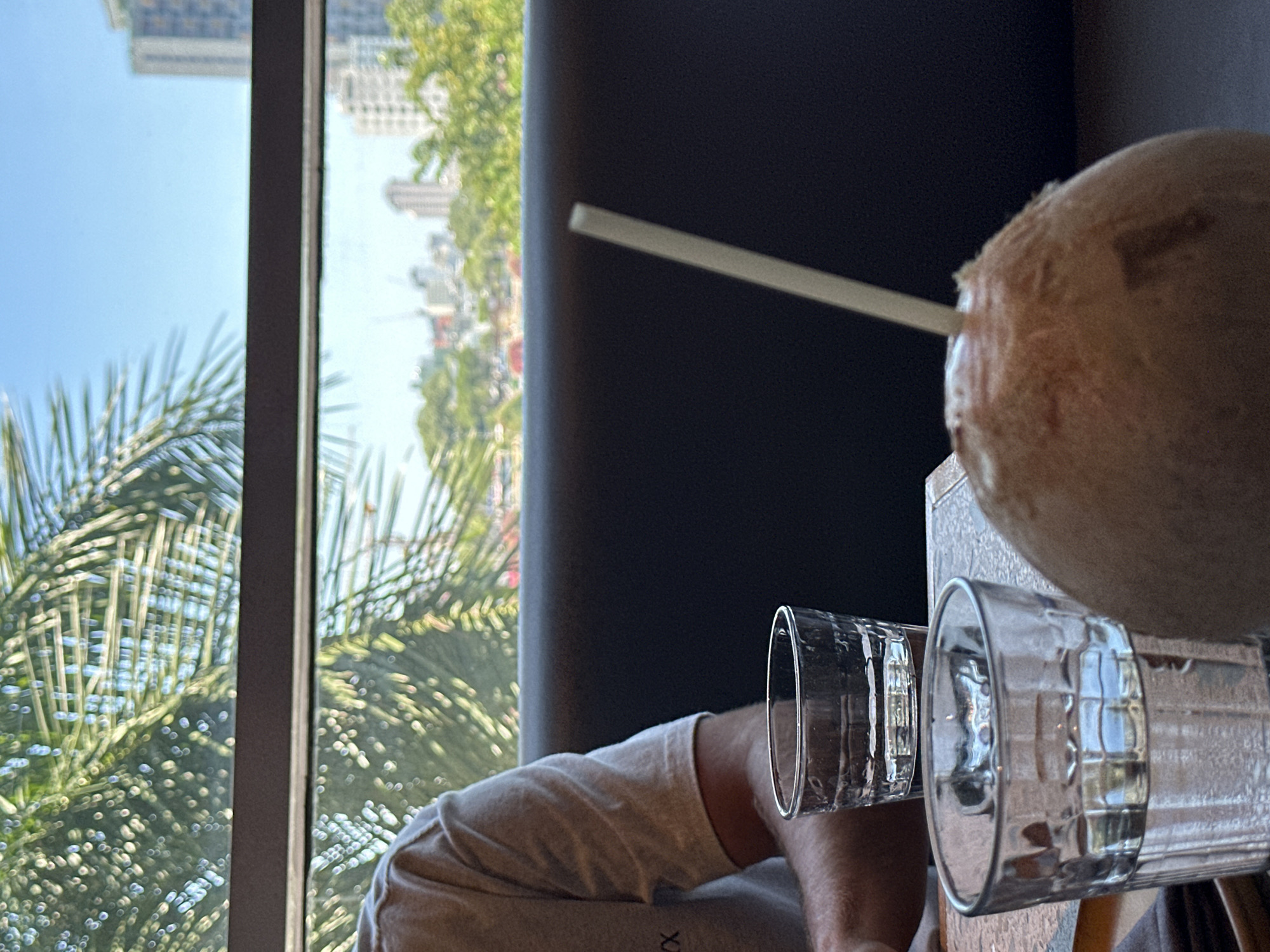 The image shows a table with two glasses of water, a whole coconut with a straw, and a person's arm and shoulder. In the background, through a window, a cityscape of Da Nang, Vietnam, can be seen, with palm trees and buildings under a clear blue sky. The scene suggests a relaxed dining or drinking setting.