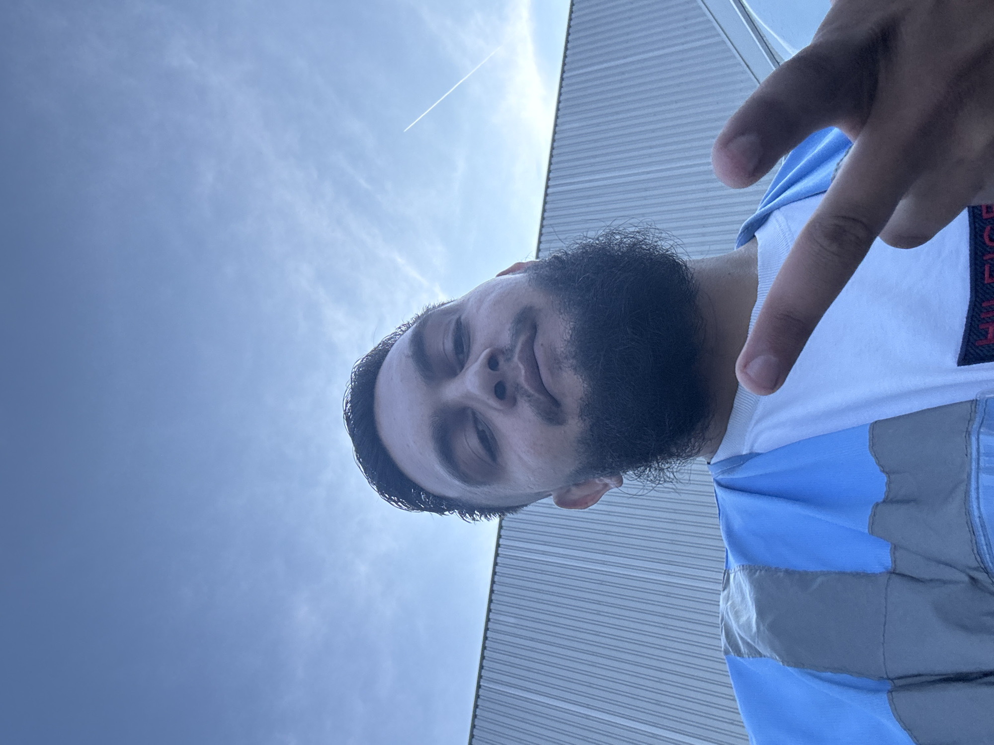 A low-angle, waist-up, selfie-style photograph features a young man with a full beard and dark hair, looking directly at the camera. He is wearing a white t-shirt and a blue and grey utility vest. His left hand is raised, with two fingers extended in a peace sign, while his right hand is blurred and out of focus. Behind him, a corrugated metal building forms the backdrop. The sky above is a pale blue with scattered white clouds and a faint contrail from an airplane. The location is the city of Boom, Belgium.