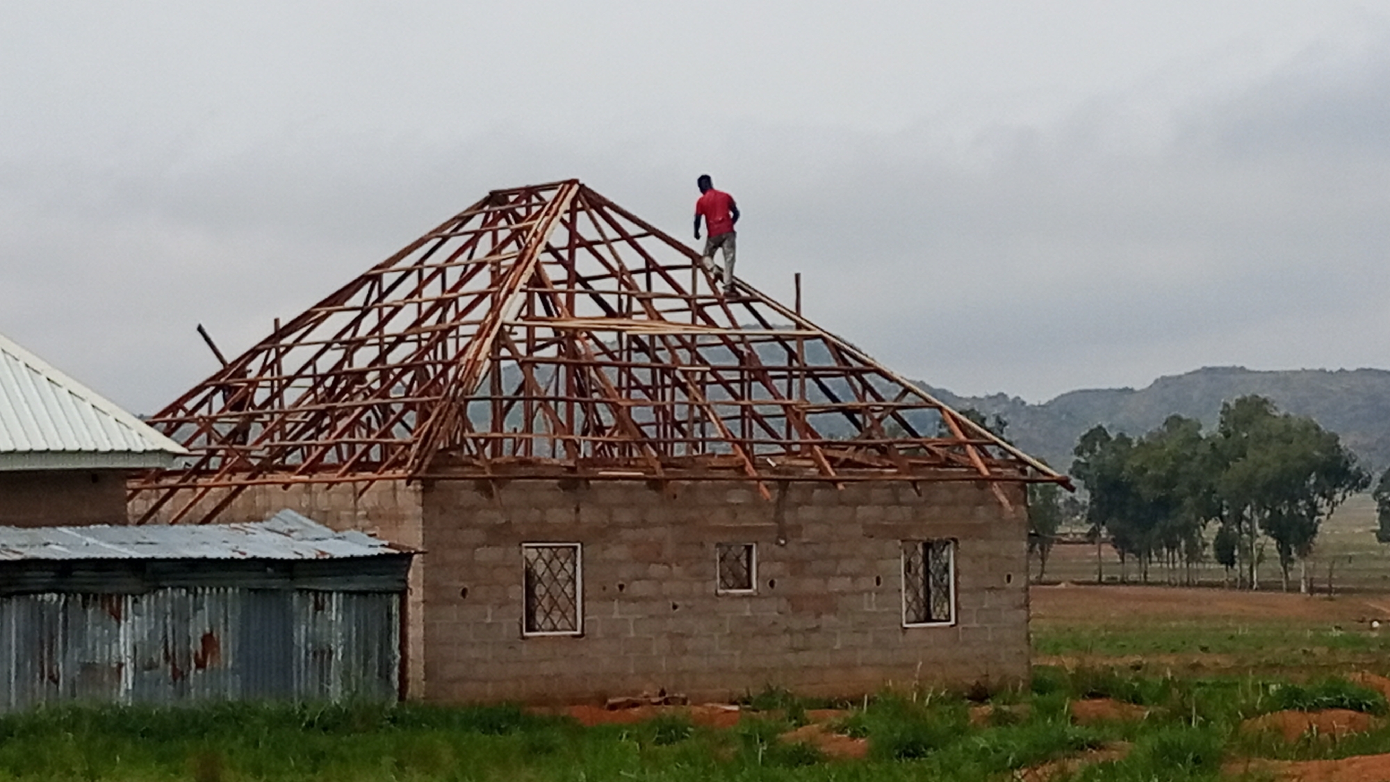 A man in a red shirt and light-colored pants is standing on the unfinished wooden frame of a peaked roof that surmounts a single-story building under construction. The building is made of light-colored concrete blocks, has several barred windows, and a partially-completed roof of wood lattice construction. To the left is another building with a white roof and corrugated metal siding. The scene is set in a grassy field in Vom, Nigeria. Beyond the buildings, trees dot the landscape, and rolling hills rise in the background under a cloudy sky.