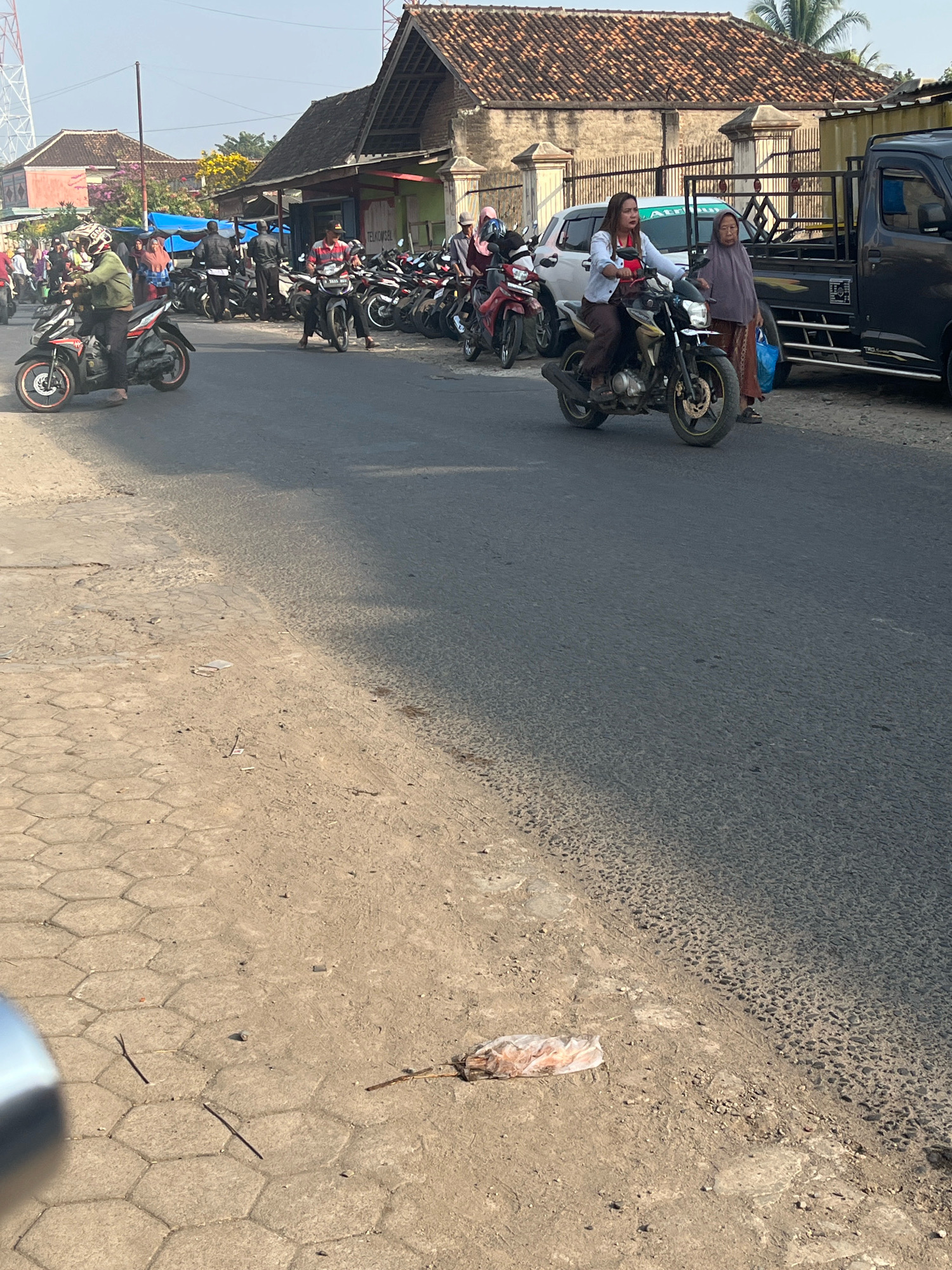 A street scene in Kemuning, Indonesia features a group of people on motorcycles. In the foreground, a person in a green jacket rides a black and red motorcycle. To the right, two women are on a motorcycle, one driving and the other a passenger wearing a purple hijab. Behind them, several other motorcycles are parked or in motion, with more people milling about. A black truck is parked on the right side of the road. Buildings with tiled roofs line the street, and trees with yellow and purple foliage are visible in the background. The ground is paved with textured concrete on the left and asphalt on the right, with some debris on the ground.