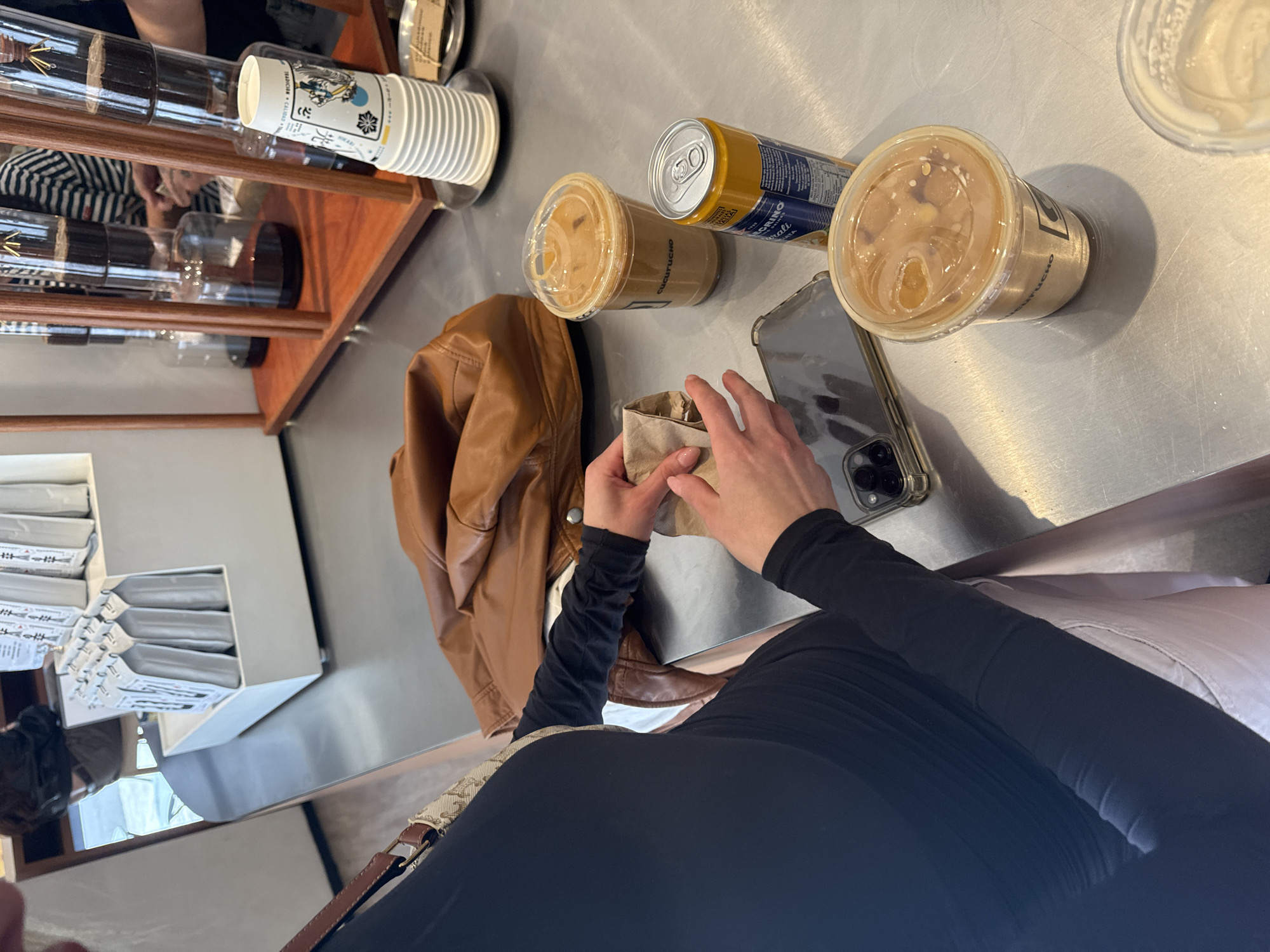 The image depicts an indoor scene at Cucurucho Café in Mexico City, Mexico. A stainless steel counter serves as the primary surface. In the foreground, a person, dressed in a black long-sleeved top and light-colored bottoms, is partially visible, holding a small brown paper bag with both hands. To their left, a brown leather jacket or bag is present, and a clear-cased smartphone with a visible camera array rests on the counter.
Arranged on the counter are three clear plastic cups containing iced coffee, one of which displays "CUCURUCHO" branding. Adjacent to one of the coffee cups is a yellow and blue can of San Pellegrino Limonata.
In the background, a wooden structure supports multiple glass cold-brew coffee towers, containing dark liquid. A stack of white paper cups, featuring a patterned design and "CUCURUCHO" text, is placed on a small metal tray. Nearby, another small metal tray holds a brown paper sachet. Further in the background, white display stands hold individually packaged white or silver coffee bags. A person wearing a horizontal striped shirt is partially visible behind the coffee equipment, and reflections suggest other figures or equipment.
