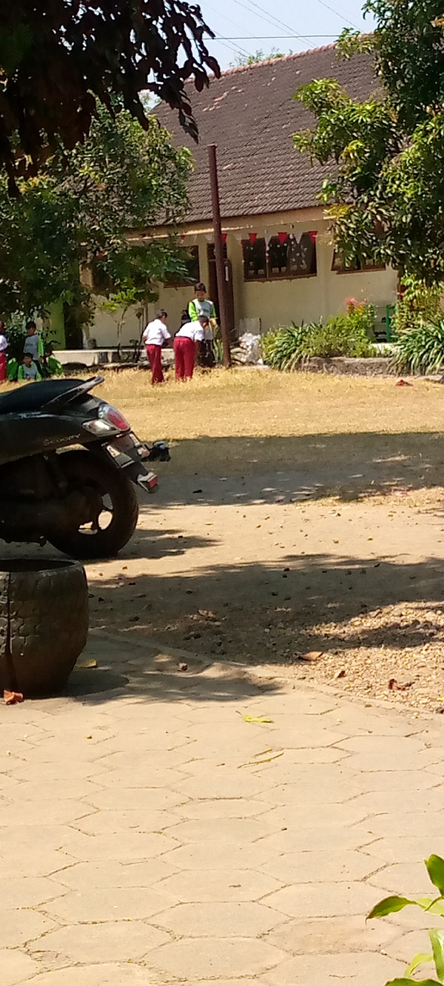 In Pacing, Indonesia, a scene unfolds in front of a building with a tiled roof. Several children in school uniforms, consisting of white shirts and red pants, are engaged in an activity on a dry, grassy area. One child stands near the building's entrance, wearing a green shirt. A dark-colored scooter is parked in the foreground, partially obscuring the view of the children. The ground in front of the scooter is paved with interlocking hexagonal tiles. The environment appears to be outdoors, with trees providing shade.