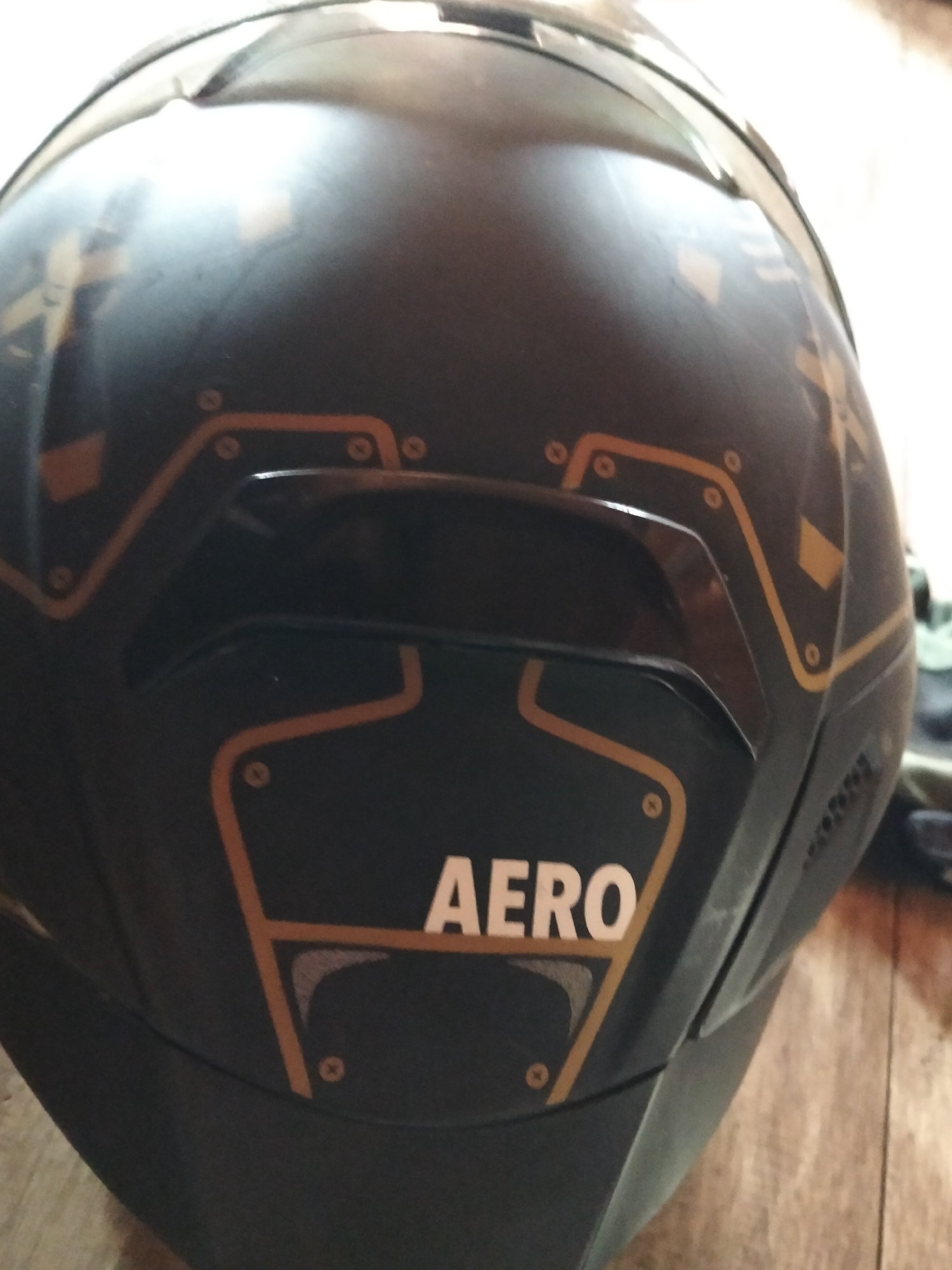 The image shows the rear portion of a dark-colored motorcycle helmet with a graphic design in a lighter color, possibly gold or yellow. The design consists of geometric shapes, including squares and lines, that resemble circuitry or paneling. The word "AERO" is prominently displayed in white capital letters on a raised section of the helmet, outlined by the same lighter color. The helmet appears to be resting on a wooden surface, possibly a floor or table, with a hint of fabric or clothing in the background on the right side. The lighting suggests an indoor environment, but there are no specific indicators of the city of Setia Jaya, Indonesia, within the visual information provided.