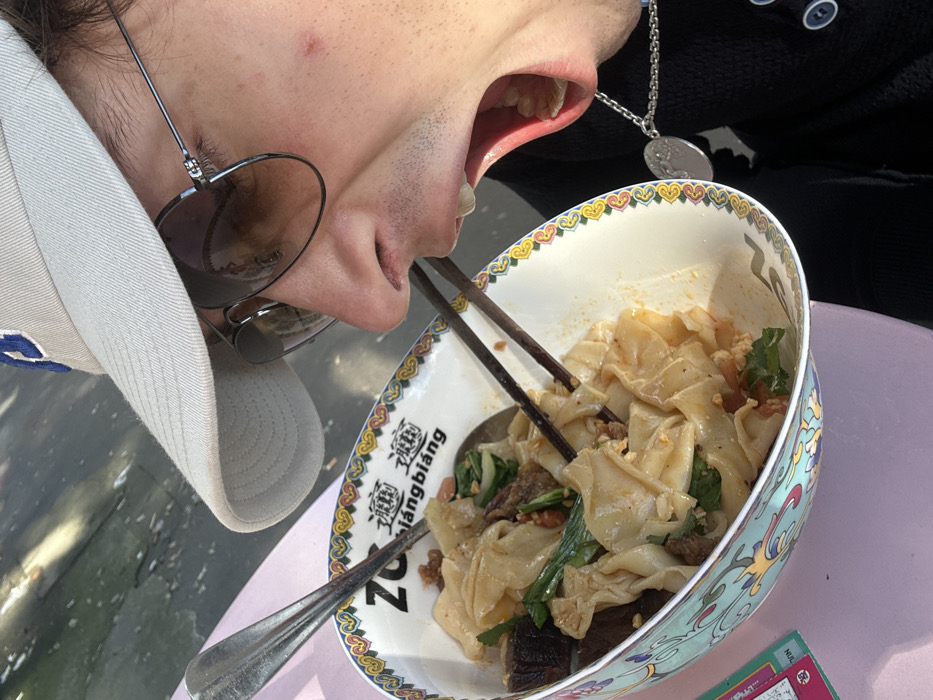 A close-up, tilted image depicts an individual preparing to eat Biangbiang noodles at the restaurant Xiang BiangBiang in Paris, France. The person, an adult, wears a light-colored baseball cap with a blue "P" logo, dark aviator sunglasses, and a dark-colored garment with buttons. A silver-colored chain necklace with a circular pendant is visible. The person's mouth is wide open, exposing the palate and teeth, as a pair of dark chopsticks emerge from a large, patterned ceramic bowl filled with broad noodles, dark meat pieces, green vegetables, and sauce. A silver spoon also rests in the bowl. The bowl sits on a light pink surface, likely an outdoor table, with an out-of-focus background showing dappled sunlight and reflections. A small red blemish is visible on the person's right cheek.