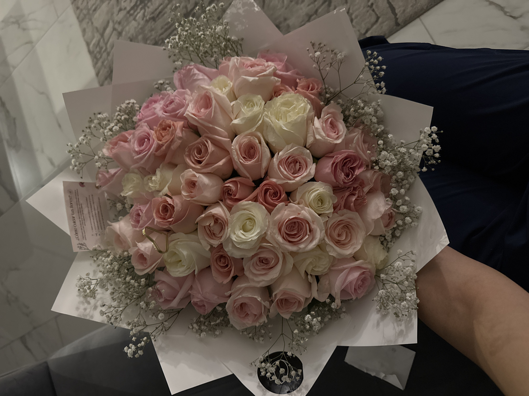 The image shows a bouquet of pink and white roses with baby's breath, wrapped in white paper. The bouquet is being held by a person wearing a dark blue garment, possibly a shirt or dress. The background includes a tiled floor and a gray, textured surface, likely a couch or rug. There is no information in the image to indicate the location is Puebla, Mexico.
