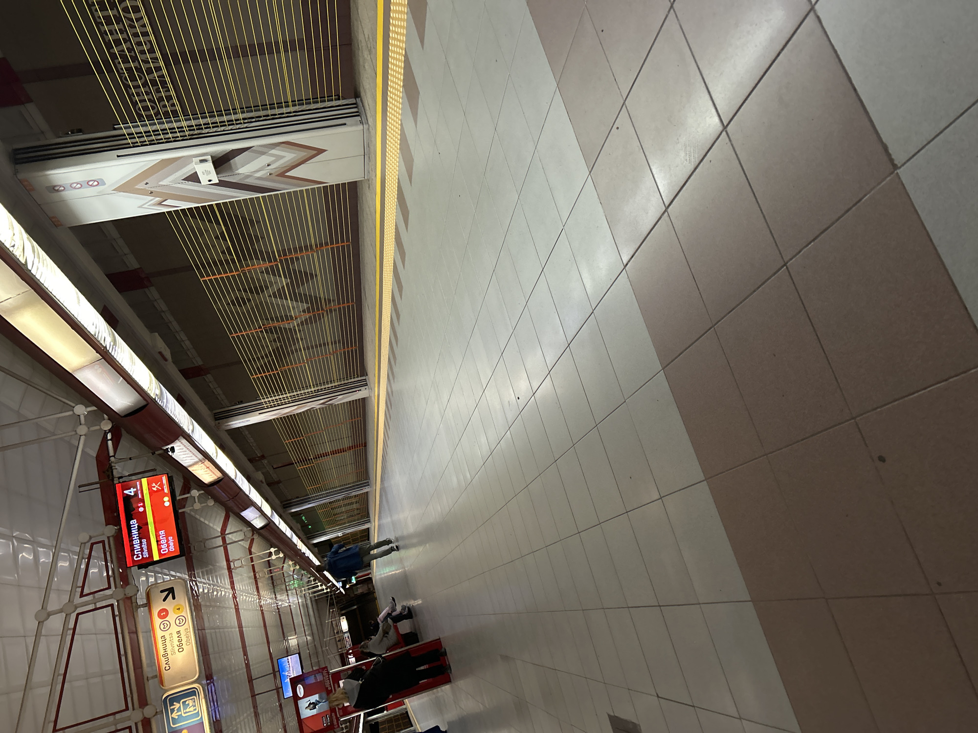 The image presents an indoor view of a modern subway station platform, identified as being in Sofia, Bulgaria. The perspective is tilted, showing the expansive platform floor dominating the right side of the frame and the station wall on the left.
The platform floor features a geometric pattern of alternating light grey and brownish-grey square tiles. A distinct yellow tactile paving strip runs along the length of the platform edge. Beyond this strip, a long, white apparatus decorated with geometric patterns in brown and beige is installed. Numerous thin, bright yellow strings extend horizontally from the top of this apparatus, forming a visible screen, likely part of an advanced safety or sensor system for the platform edge.
On the station wall to the left, which is tiled in white with a red-brown grid pattern and horizontal accents, several information signs are displayed. A prominent red digital screen clearly shows "СЛИВНИЦА" (Slivnitsa) and "ОБЕЛЯ" (Obelya) in both Cyrillic and Latin script, alongside a large number "4", indicating the subway line. An arrow and a train icon are also visible. Below this, a yellowish-brown sign also lists "СЛИВНИЦА" (Slivnitsa) and "ОБЕЛЯ" (Obelya) with a right-pointing arrow and a small smiling face icon.
Several people are scattered across the platform, either standing or walking, suggesting they are waiting for a train or transiting through the station. Their activities are typical of a public transport environment. The station is brightly illuminated by artificial overhead lighting. The clean design and legible signs in both Cyrillic and Latin characters align with the provided context of Sofia, Bulgaria. No specific time of day or weather can be discerned from the indoor setting.