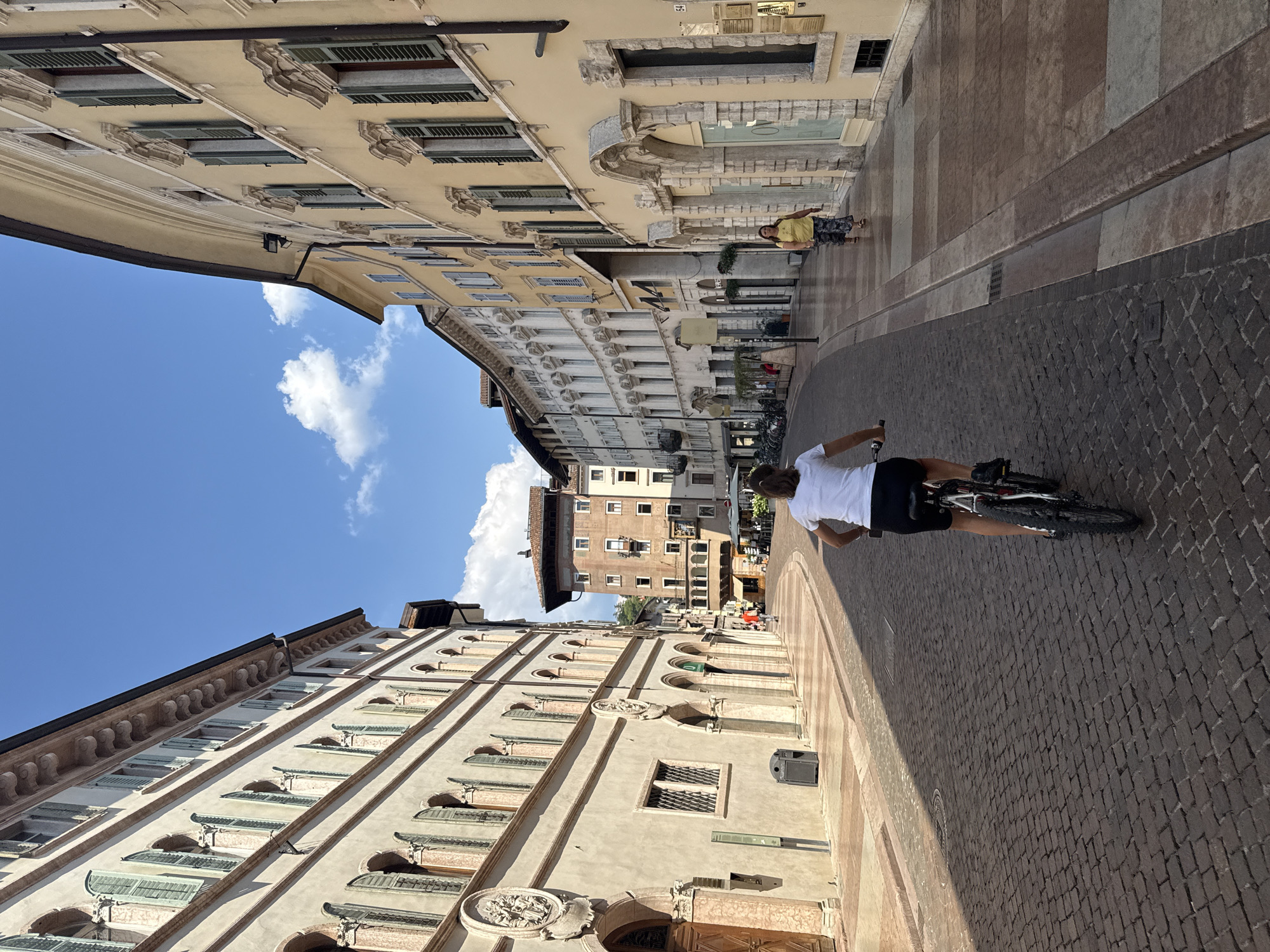 In Trento, Italy, a woman is biking down a narrow, cobblestone street lined with historic buildings. The architecture suggests a European city center, with multi-story buildings featuring arched windows and ornate facades. The buildings on the left are a pale beige, while those on the right are a warm yellow. The street is paved with dark grey cobblestones and a lighter colored sidewalk runs along the right side of the street, made of alternating light brown and grey tiles. The sky is bright blue with a few scattered white clouds. The woman, wearing a white t-shirt and black shorts, is the primary subject, seen from behind as she cycles away from the camera. Another woman in a yellow top and patterned pants walks on the sidewalk in the middle distance. The street is relatively empty, suggesting it might be a quiet time of day or a less-trafficked area. The overall scene conveys a sense of everyday life in an old European city.