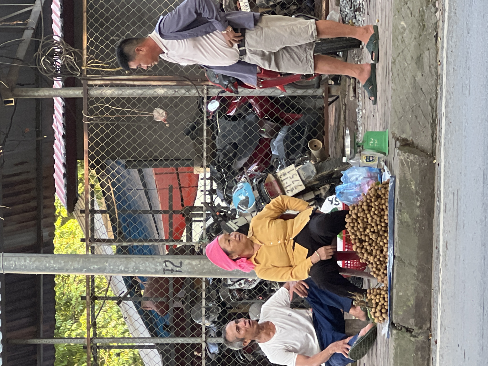 The image captures a street scene in Liên Sơn, Vietnam. In the foreground, two individuals are seated on a curb, with a substantial pile of longan fruit displayed on a blue tarp. A small green weighing scale is placed next to the fruit. A third individual, a man, stands to the right, facing the seated pair. The background features a chain-link fence, behind which numerous motorcycles are parked. Overhead, a corrugated metal roof structure is visible. The setting appears to be an outdoor market or roadside stall.