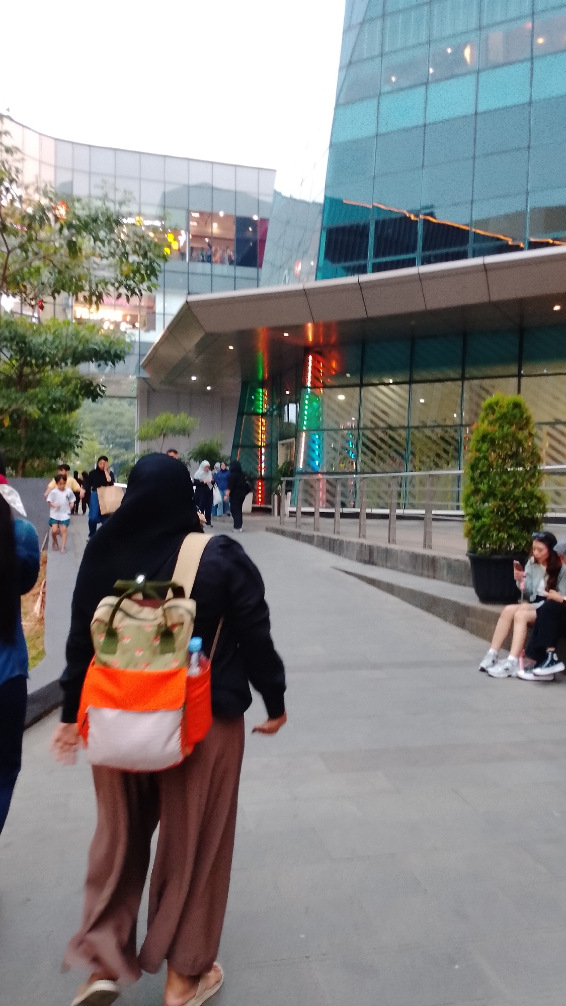 A medium shot captures the rear view of a person wearing a black hijab and loose brown pants, walking away from the camera on a paved walkway in front of a modern building. They are carrying a backpack with orange, beige, and green panels. To the right, two women are seated on a raised planter, one looking at her phone. In the background, other individuals, including children, walk along the walkway and near the building's entrance. The building features a glass facade with diagonal lines and colorful vertical lighting. Trees and foliage are visible to the left. The overall scene appears to be in an urban, commercial area, potentially a mall or a modern office complex, located in South Tangerang, Indonesia. The lighting suggests it is either early morning or late afternoon.