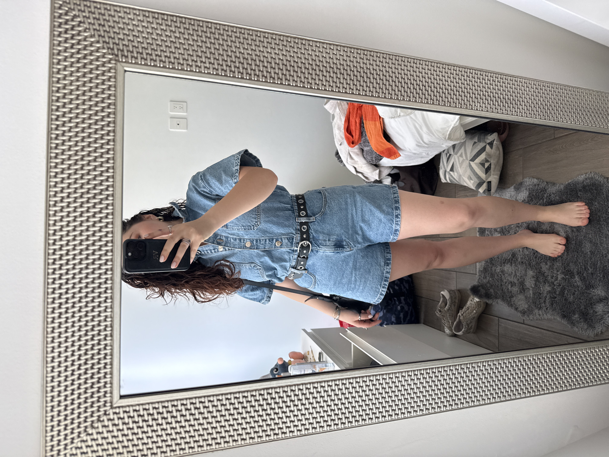 A young woman takes a mirror selfie indoors. She wears a denim romper with a black belt and holds a black smartphone. Her hair is curly and brown. The mirror has a textured, silver frame. Reflected in the mirror is a cluttered room, with items on a shelf and clothes piled on the floor. A gray rug lies on the floor. The location is possibly in Mexico City, Mexico, but there is no definitive proof.