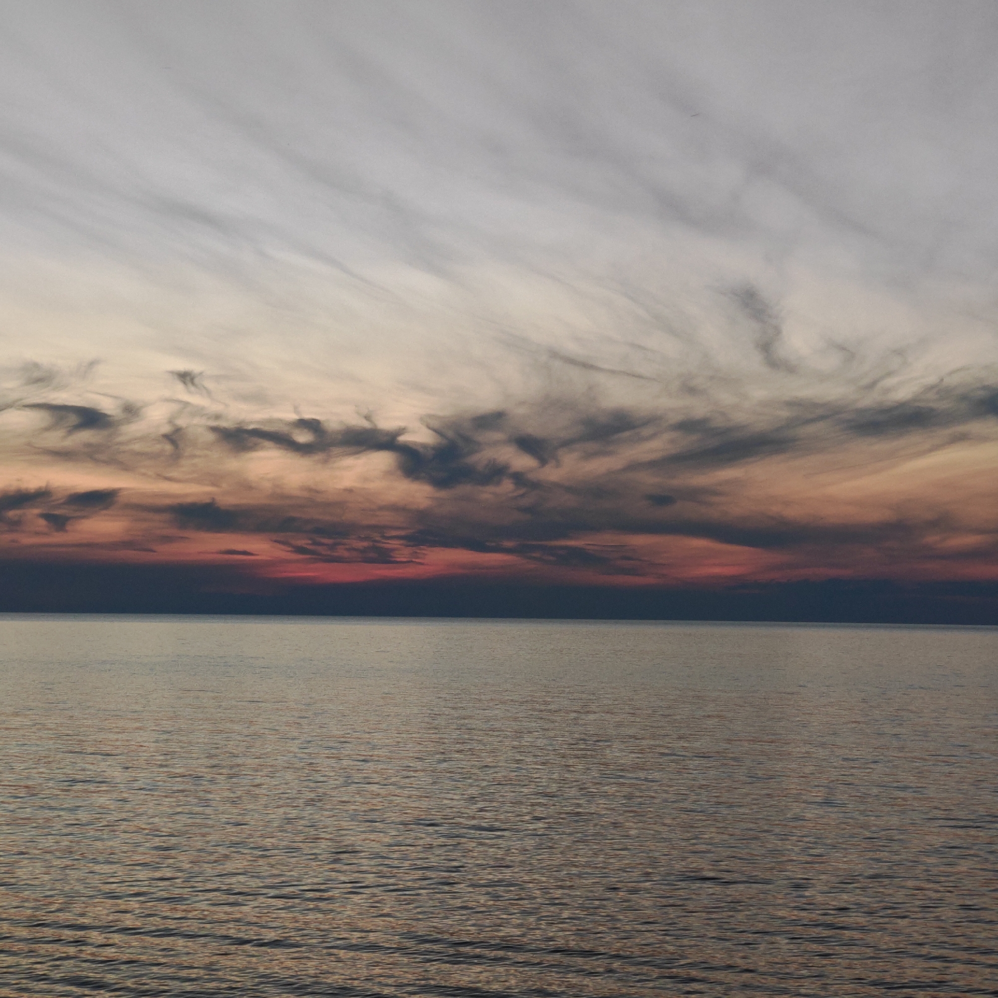 A vertical photograph captures an open body of water at sunset in городское поселение Щелкино, Ukraine. The sky occupies the upper two-thirds of the frame, exhibiting a distinct color gradient and cloud formations. The uppermost section features a pale grey, slightly textured sky with faint, wispy cloud streaks. A minute dark speck, possibly a distant aircraft or bird, is barely discernible in the upper left quadrant. Below this, a band of more defined, elongated, and somewhat swirling clouds extends across the middle of the sky. These clouds transition in color from light orange and pale yellow at their top edges to progressively deeper shades of brick red, purplish-grey, and dark navy closer to the horizon line. The lowest segment of the sky, directly above the water, is a dark, intense purplish-red. The horizon line separating sky and water is sharp and extends horizontally across the image. The lower third of the image depicts the water surface, which is relatively calm with subtle, widespread ripples. The water reflects muted, warm tones from the sky, appearing as a blend of coppery browns and deeper blues or greys. There are no visible landmasses, structures, vessels, or individuals present within the scene.