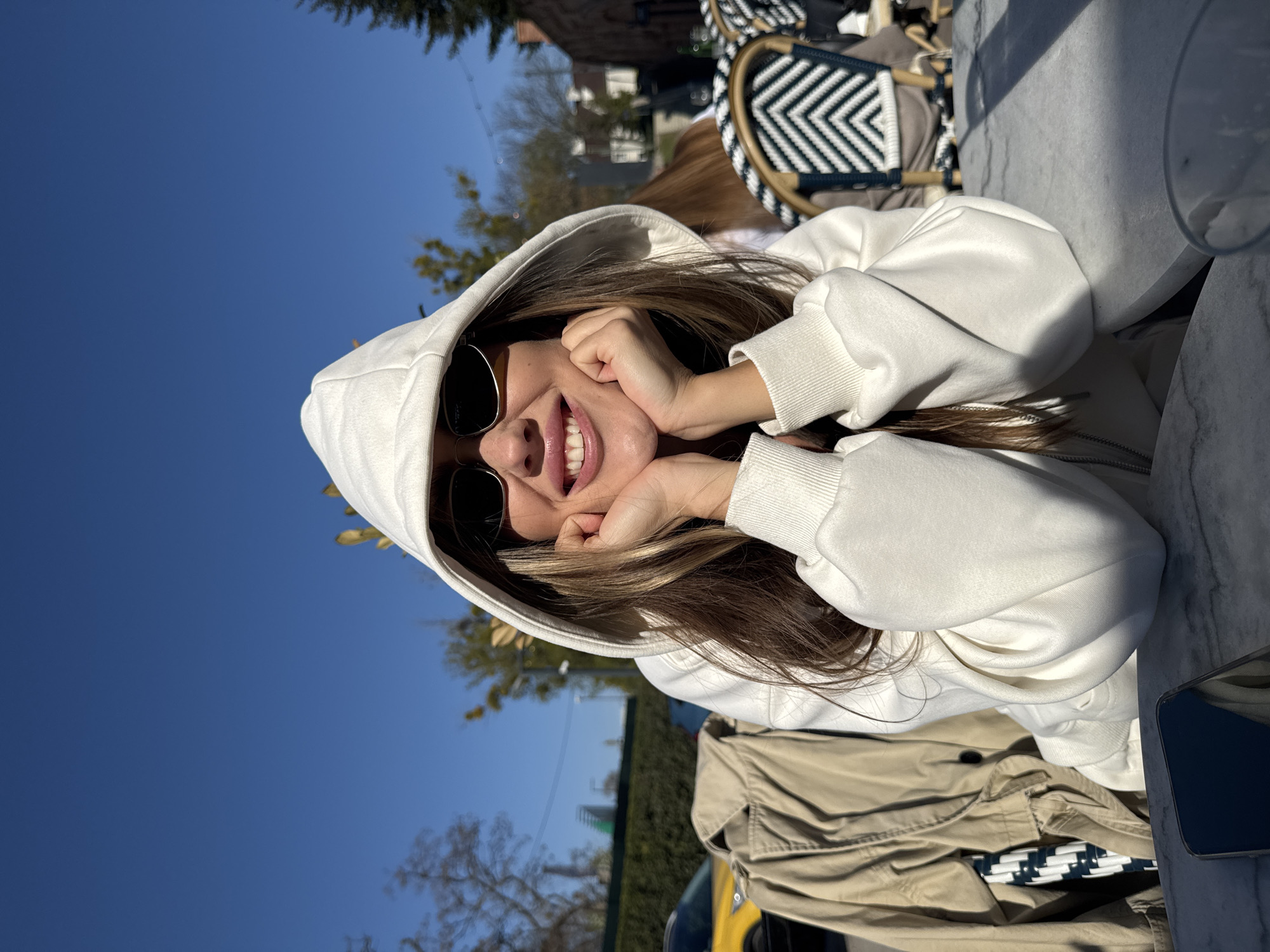 A smiling young woman wearing a white hoodie and sunglasses is outdoors with her hands on her cheeks. She has long brown hair with blonde highlights. The photo appears to be taken from a low angle, looking up at the woman. The background shows trees, blue sky, and buildings. A table with a marble top is visible in the lower right corner, along with a jacket. The presence of a distinctive black and white patterned chair back suggests the image may have been taken at a cafe or outdoor seating area. While it is not possible to confirm from the image alone, the general style and vegetation are consistent with typical outdoor seating areas in Warsaw, Poland.