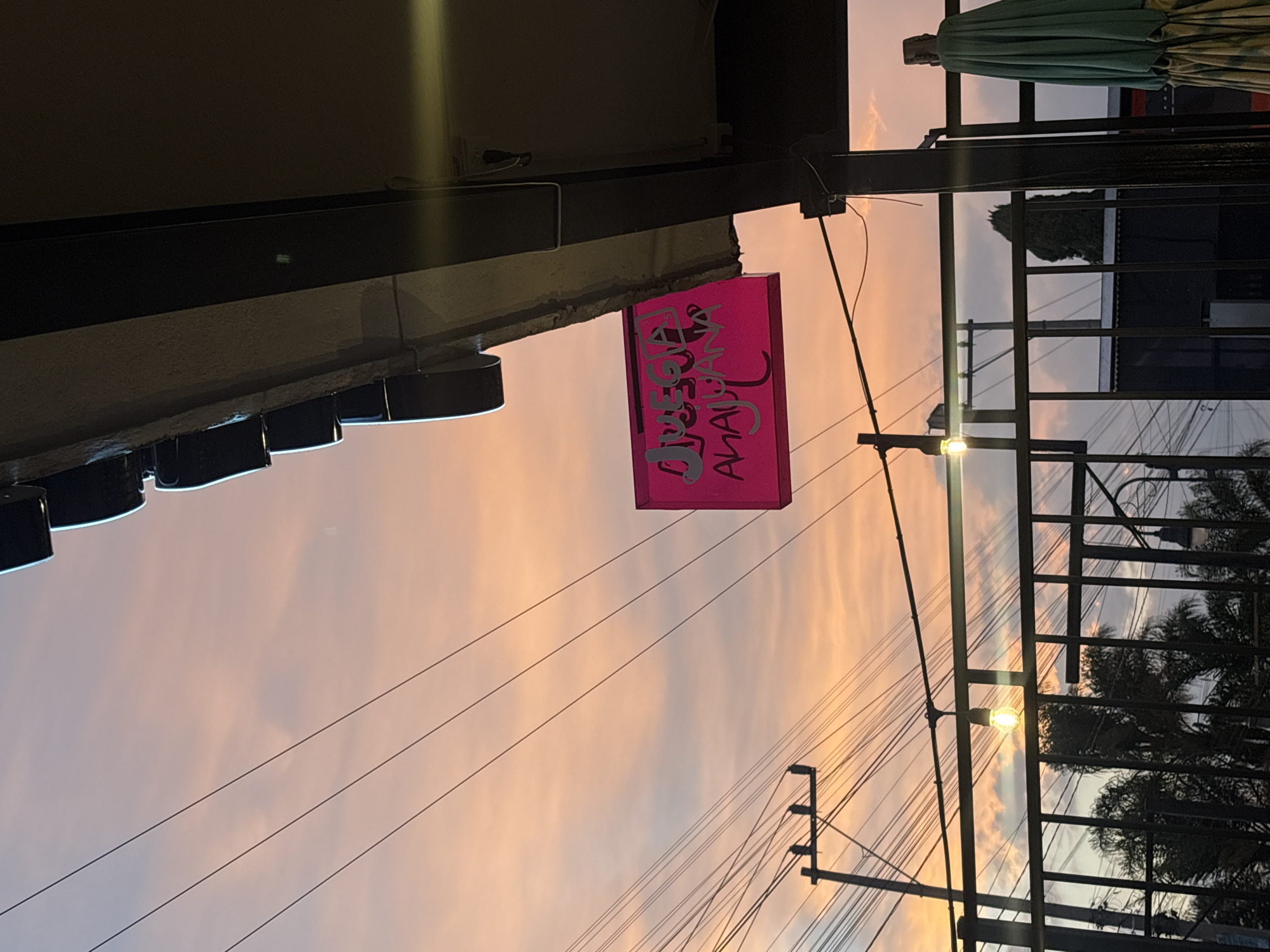 Captured from a low angle, the photograph presents an urban scene at dusk in San Andrés Cholula, Mexico. The sky, painted with hues of orange and soft pink, forms the backdrop. Overhead power lines crisscross the sky.
In the foreground, architectural elements of a building structure are visible, along with the edge of a dark green patio umbrella. A rectangular, pink sign reading "JUEGOS AHAUANA" hangs suspended from the building. The sign appears to be illuminated.
A black metal fence-like structure stretches across the right side of the frame. Behind it, silhouettes of palm trees and the façade of another building can be seen. A street lamp attached to the fence structure emits a bright glow.