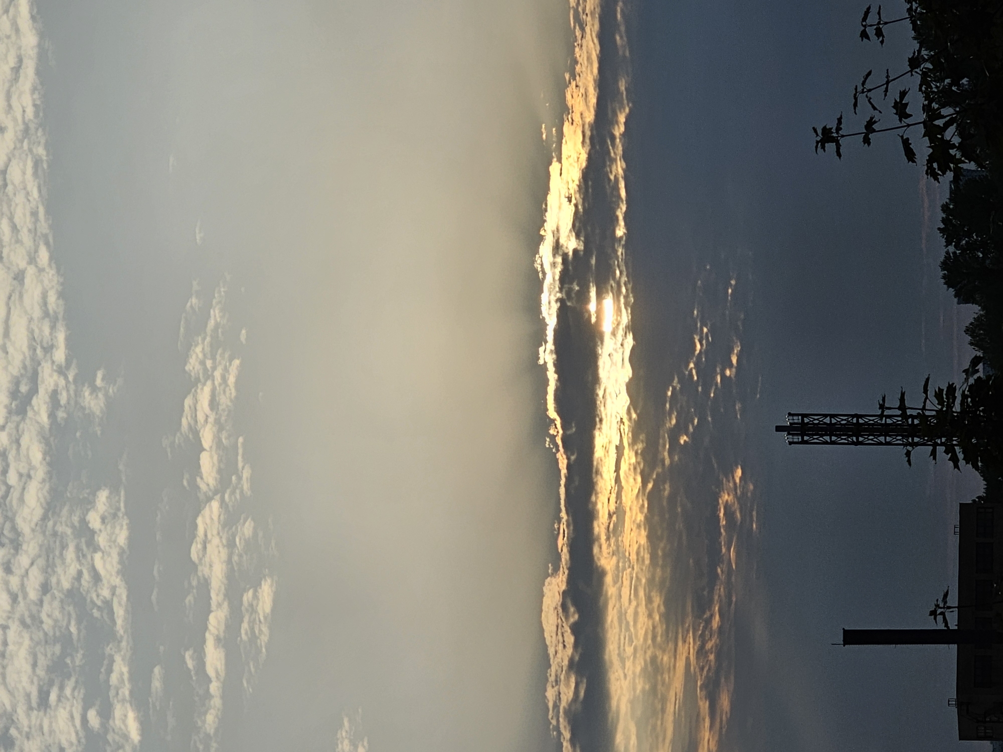 A vertical, outdoor, eye-level shot captures a sky filled with clouds at sunset. The sun is partially obscured by a band of dark clouds, with rays of light emanating through the gaps, illuminating the undersides of the clouds with a warm, golden hue. In the foreground, silhouetted against the sky, are several structures. To the left, a tall, dark chimney rises from a low-rise building. To the right of the chimney is a lattice tower, also silhouetted. Further to the right, the dark silhouettes of trees are visible. The overall impression is a moody, atmospheric cityscape at dusk. This image was taken in Khimki, Russia.