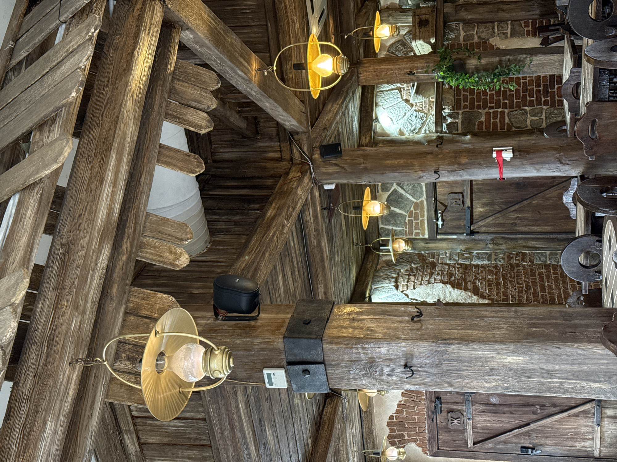 The image displays the unoccupied interior of a rustic-themed restaurant or pub, characterized by extensive use of dark, rough-hewn wood for the ceiling beams, planks, and large structural pillars. Walls feature a combination of exposed brickwork and natural stone. Multiple brass-colored electric pendant lights, styled as antique oil lanterns, hang from the wooden ceiling, illuminating the space with a warm, yellowish glow.
Prominent features include massive wooden pillars. One central pillar contains a small white digital display (likely a thermostat or hygrometer) and multiple black metal coat hooks. Two black wall-mounted speakers are visible, one affixed to a pillar, the other to a ceiling beam. Two substantial wooden plank doors are present; the door on the left bears a Cyrillic character "Ж" (Zhe), indicating a women's restroom, while the door on the right has "М" (Em), denoting a men's restroom. Both doors feature traditional iron hardware. Dark wooden tables and chairs, some with circular backs, are arranged throughout the dining area. White air ducting is visible running beneath some ceiling elements. A small artificial green plant hangs from a wooden beam. A small wooden chest rests on a high ledge near the 'M' door. The scene is static and devoid of individuals, capturing the establishment's interior located in Moscow, Russia.