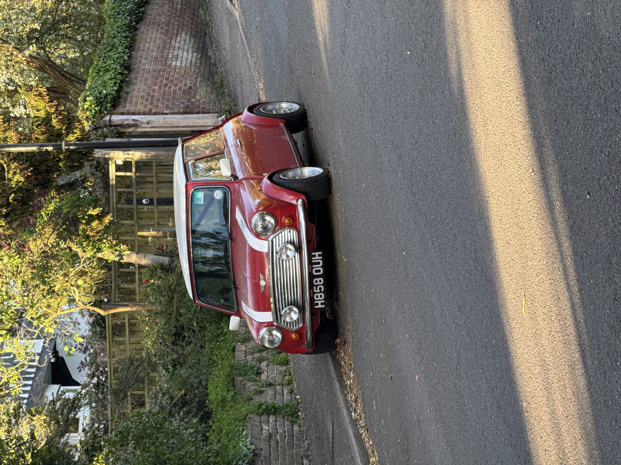 A red Mini Cooper with a white roof and racing stripe is parked on the side of a paved road. The car's license plate reads "H858 OUH". The surrounding environment includes a brick wall, various trees, bushes, a wooden fence, and a house with a black and white awning. The location is London, United Kingdom.