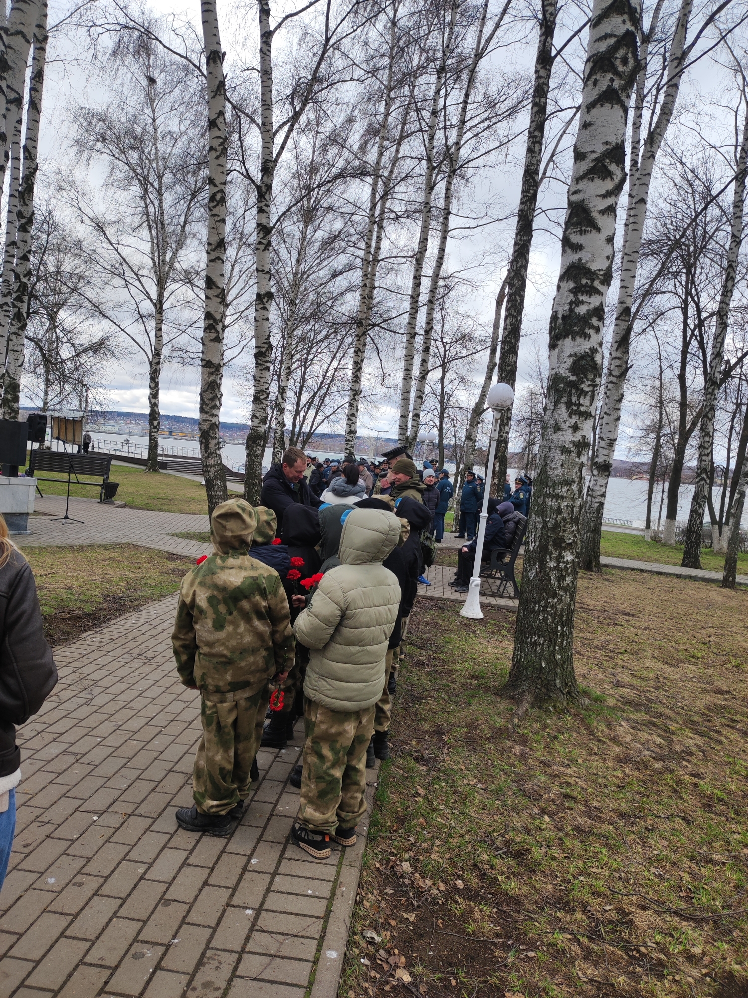 The scene is an outdoor gathering of people at what appears to be a memorial or event in Votkinsk, Russia. Several individuals, including children dressed in camouflage attire, are holding red flowers. A group of people stands on a paved walkway, while others are gathered around a monument and benches in a park-like setting. Birch trees with distinctive white bark are prevalent. In the background, there is a body of water. A lamp post and audio equipment are also visible.