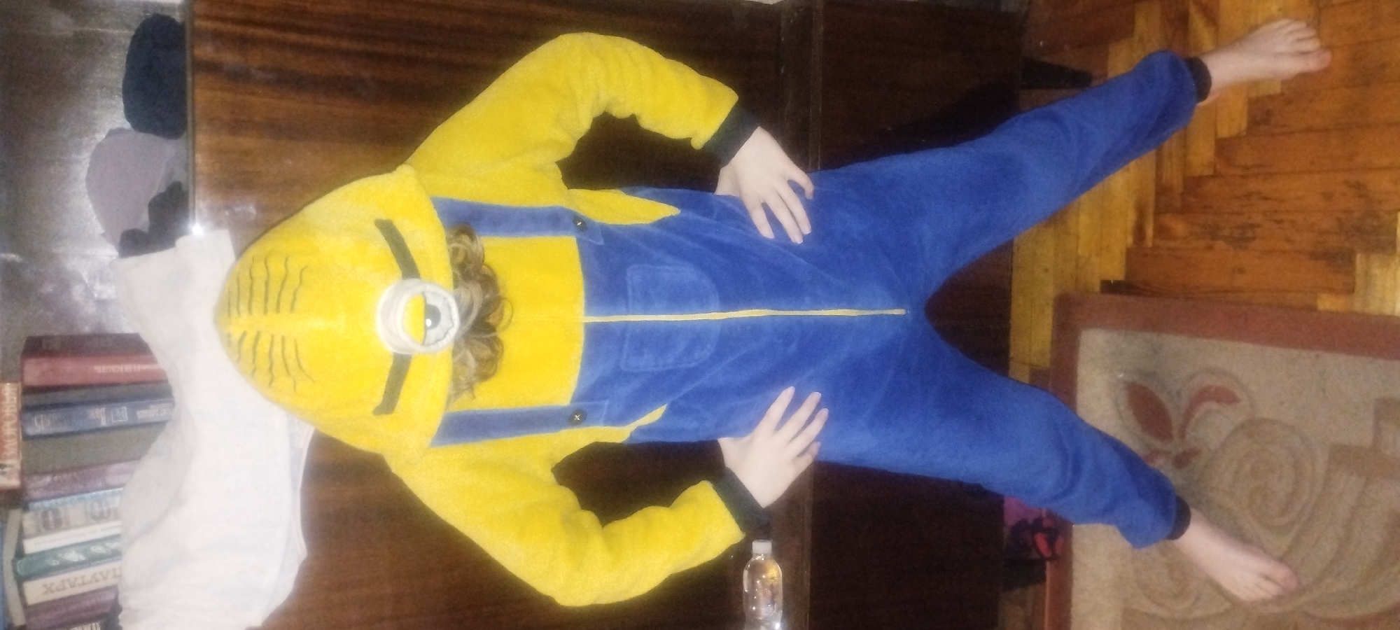 An individual is standing in a room, wearing a yellow and blue minion costume. The costume covers their entire body, including a hood with a single eye and black stripes, and blue overalls with yellow trim and black buttons. Their hands are on their hips. The individual is barefoot and standing on a rug with a floral pattern. Behind them is a wooden dresser and a shelf filled with books. The floor appears to be made of wood. The lighting is dim.