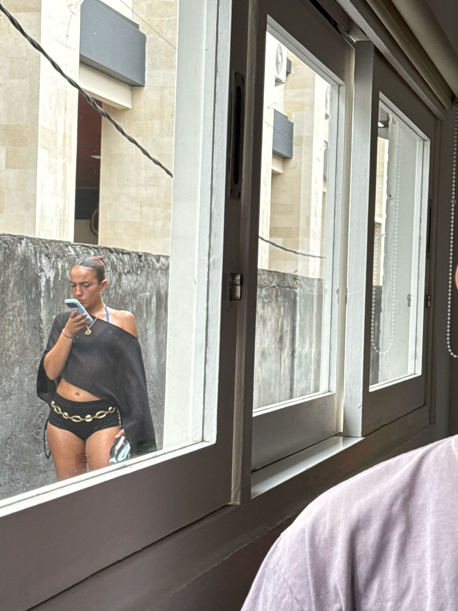 A young woman with dark hair pulled into a bun is standing outside, looking at her phone. She is wearing a black off-the-shoulder top, black shorts, and a gold chain belt. She is holding a light blue smartphone in her right hand. In the background, there is a grey concrete wall and a building with light-colored stone. The scene is viewed through a window, with the reflection of a person wearing a purple shirt visible in the foreground. The window frame is white, and there are vertical blinds on the right side.