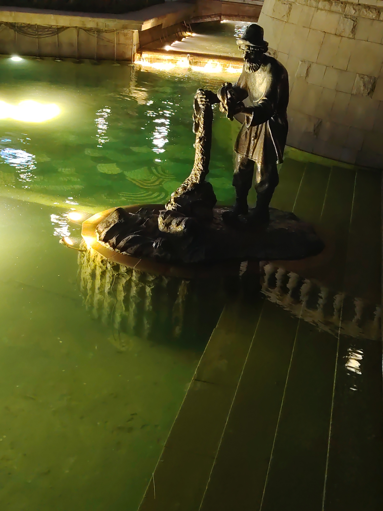 A bronze statue of a man wearing a hat and beard stands in a murky green fountain at night. He is holding a rope or chain. The statue is on a rock-like pedestal. The fountain is lit from below, casting an eerie glow on the water and statue. The background shows a stone wall and more lighting. The water is reflective, showing the lights and the statue's shadow.