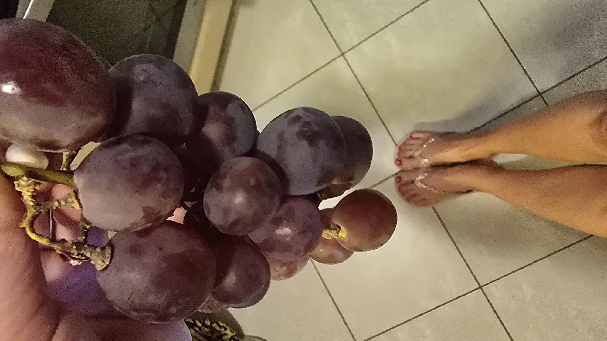 An indoor, top-down shot features a person's hand holding a large bunch of dark red or purple grapes in the foreground. The grapes appear ripe, with some individual berries showing slight variations in color and a few stems visible. In the background, the lower legs and feet of a person are visible on a light-colored tiled floor. The person is wearing light-colored flip-flops, which have decorative, chain-like straps extending over the top of the foot and between the toes, resembling toe rings. The toenails are painted a bright red color. The floor consists of large, light-toned square tiles with darker grout lines, suggesting a domestic setting like a kitchen or bathroom. The lighting appears to be artificial and internal, casting soft shadows. No specific time of day, weather, or emotions can be discerned, and there is no visible text in the image.