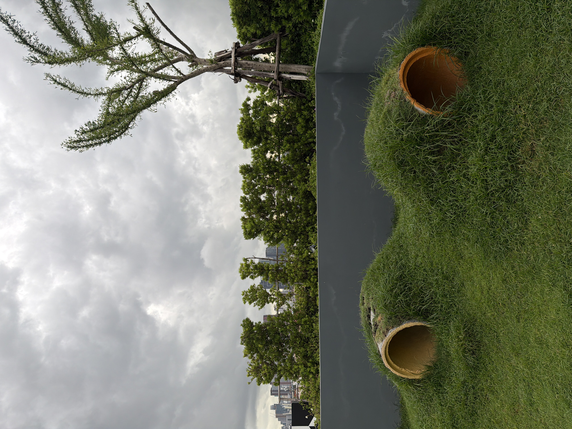The image captures a rooftop scene in Bangkok, Thailand, under a cloudy sky. In the foreground, two large, orange, cylindrical pipes are partially buried in mounds of lush green grass. These pipes appear to be part of a playful landscape feature, possibly for children or pets, with their openings visible and covered in grass. A low, dark gray wall runs across the middle ground, separating the grassy area from a line of manicured green trees and shrubs behind it. Beyond the greenery, the cityscape of Bangkok is visible in the distance, with various buildings and skyscrapers under the overcast sky. To the right, a single tree with a distinctive, somewhat sparse canopy is supported by wooden stakes. The overall impression is a blend of urban greenery and a whimsical outdoor play area situated high above the city.