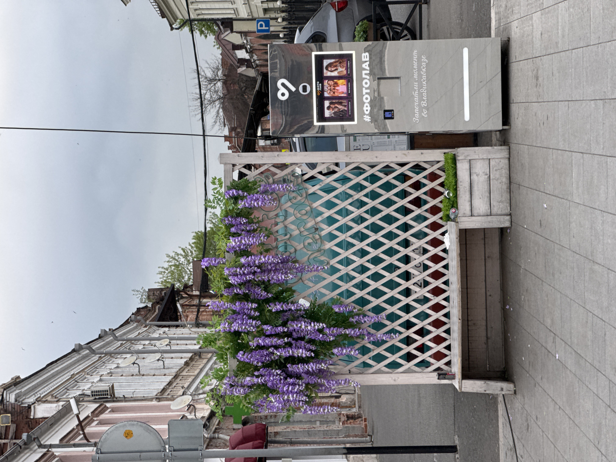 A photo booth stands on a paved street in front of a weathered building. The building's facade is brick, with layers of window frames and a sloping roof. A street sign is partially visible on the left. In front of the photo booth is a wooden structure, possibly a bench with a lattice back. The lattice is adorned with vibrant purple wisteria. A car is parked behind the photo booth. The sky is overcast. The photo booth is labeled "#ФОТОЛАВ" and includes Russian text mentioning Vladikavkaz. Based on the metadata, the location is identified as Akhieli, Georgia.