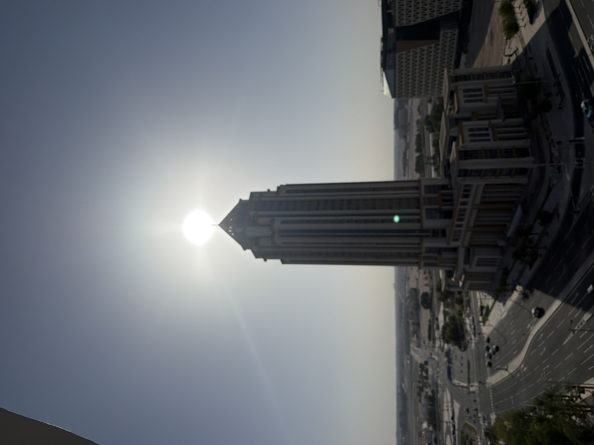 An elevated, slightly blurred photograph captures a prominent skyscraper silhouetted against a bright, high sun in an unknown city, unknown country. The sun, centered directly behind the building's apex, creates a strong starburst lens flare and significantly overexposes the upper sky, rendering it a brilliant white. The rest of the sky is a pale, hazy blue-grey.
The central subject is a tall, multi-story building exhibiting a classical or Art Deco-inspired architectural style, characterized by strong vertical lines, recessed window sections, and a distinctive pointed, faceted cap. Due to the backlighting, its facade appears dark, likely an off-white or light stone, with some glints of reflection.
Below and around the skyscraper, a complex network of multi-lane roads with white markings is visible. Several vehicles, primarily cars, are present on the roadways, some appearing to be in motion. To the right of the main building, a smaller, similarly styled structure is visible. Further right, a distinct, multi-story building with a dark, patterned facade featuring geometric or lattice-like designs stands alongside the road. Sparse green vegetation, including what appear to be palm trees, is distributed along the road shoulders and near buildings. Shadows from the buildings and trees are cast towards the left, indicating a high sun angle.
The background reveals a flat, arid-looking landscape extending to a hazy horizon, with scattered low-rise structures and roads, suggesting a sprawling urban or suburban environment. The overall scene is captured from a high vantage point, possibly a higher floor of another building, under strong, direct sunlight.