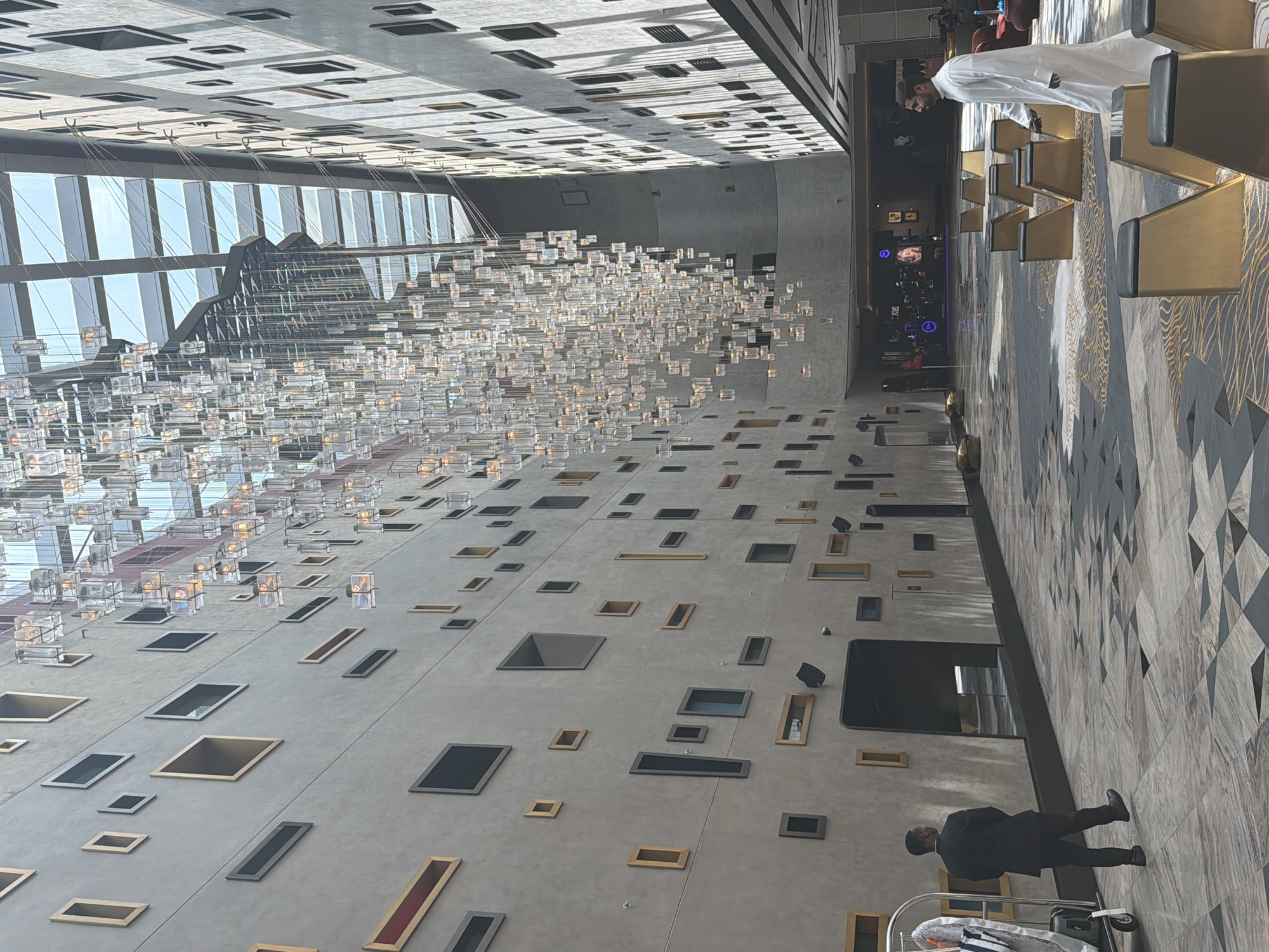 This image shows the interior of a modern hotel lobby in Dubai, United Arab Emirates. The lobby features a tall, open atrium with a skylight and a large, elaborate chandelier made of numerous small glass elements hanging from the ceiling. The walls are textured and adorned with many irregularly placed rectangular openings of varying sizes and colors, some appearing as windows or decorative elements. The floor is covered with a patterned carpet in shades of gray, black, and gold. In the foreground, a man in a black suit walks away from the camera, pushing a luggage cart. Further into the lobby, a man in traditional white attire walks towards the camera. Seating areas with gold-colored chairs with black cushions are visible. In the background, there is a large screen displaying images and some seating areas. The overall impression is of a luxurious and contemporary space.