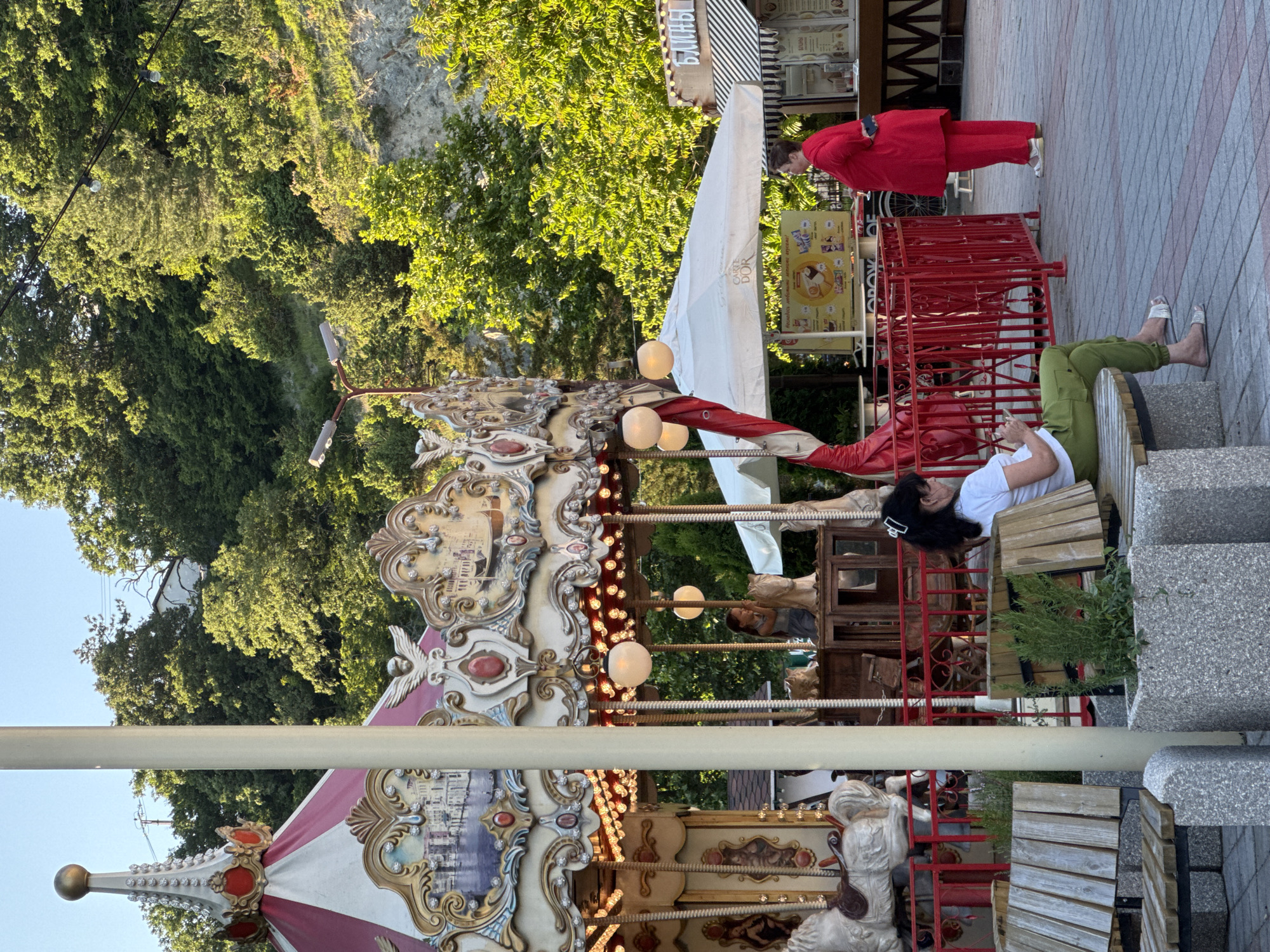 In the city of Abrau-Dyurso, Russia, a carousel is set up outdoors, a prominent feature in the scene. The carousel is ornate, adorned with red and white striped fabric, intricate gold detailing, and what appear to be painted murals depicting a cityscape. Several wooden horses are visible, some with decorative saddles. A young girl is seen on the carousel, appearing to be one of the few passengers.
In the foreground, a woman with dark hair, wearing a white t-shirt and green pants, sits on a wooden bench, looking at her phone. She is barefoot, wearing white sandals. To her right, another woman, dressed in a bright red suit, stands looking to her left. She holds a glass in her right hand and has her left hand on her hip.
Behind the carousel, a white umbrella tent with "Carte D'Or" branding is set up, suggesting it may be a concession or rest area. Adjacent to it, a food stand with a striped awning is visible. The background consists of a lush green hillside covered in trees, with rocky outcrops. The ground is paved with interlocking bricks. The lighting suggests it is late afternoon or early evening.