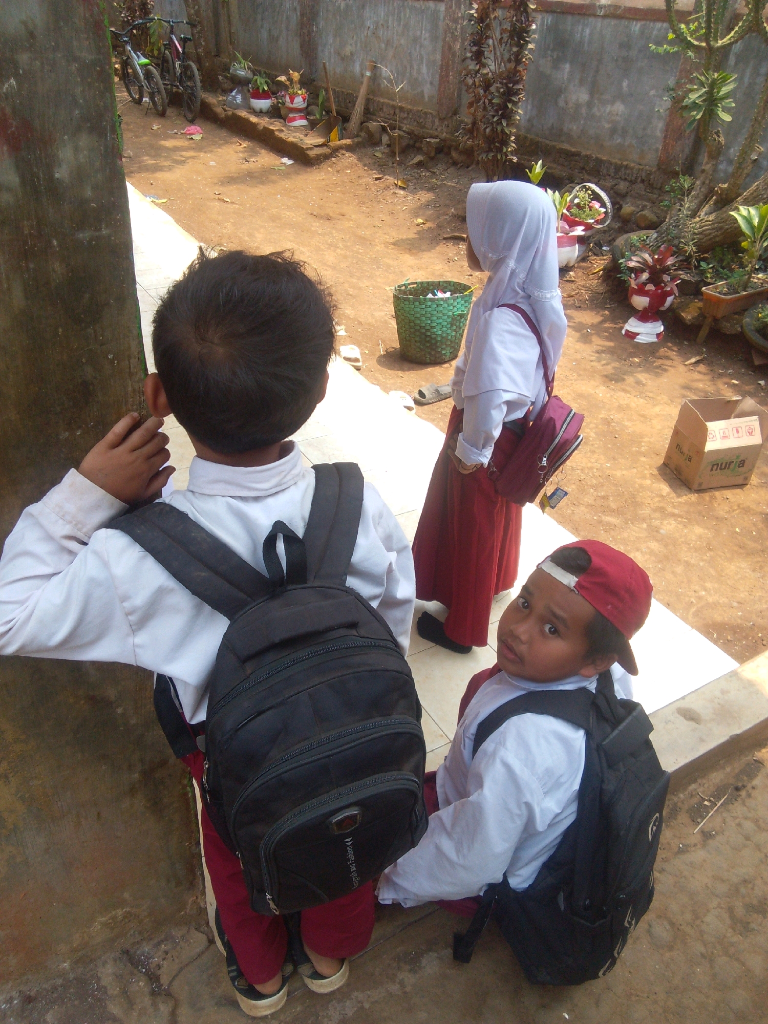 In Betek, Indonesia, two children wearing school uniforms are in an outdoor courtyard with a concrete pathway. The child on the left, seen from behind, wears a white long-sleeved shirt and maroon pants, carrying a black backpack. The child on the right, facing forward, wears a white long-sleeved shirt, maroon pants, and a red baseball cap. This child also carries a black backpack. Behind the children, a girl in a white hijab and maroon skirt stands with a maroon backpack. The environment includes a textured concrete wall, various potted plants, two bicycles, and a cardboard box. The ground is a mixture of dirt and concrete.