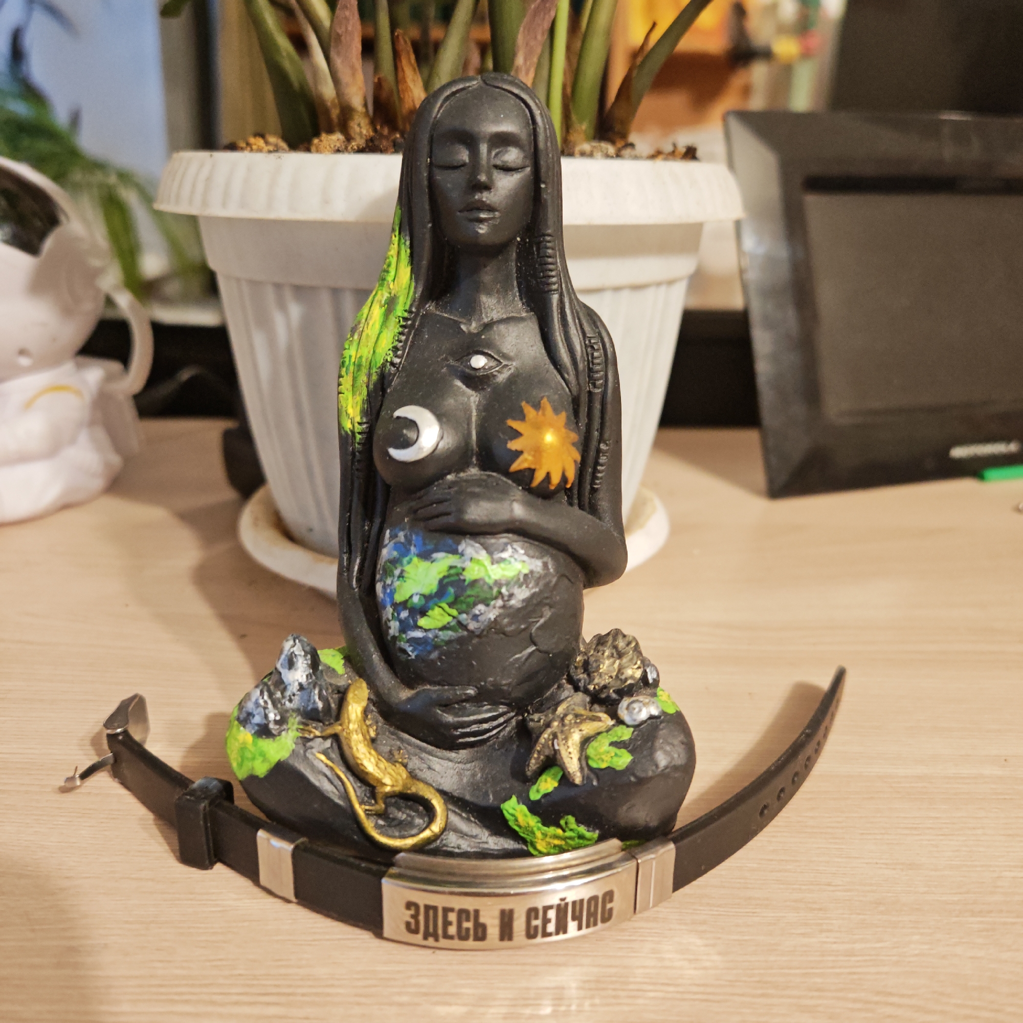 The image features a statue of a pregnant woman holding a globe, with a crescent moon on one breast and a sun on the other. The statue is black with green highlights in the hair and on the base. It is adorned with a third eye on her forehead. The base features a golden lizard and other small creatures. A black leather strap with a silver plate bearing the inscription "ЗДЕСЬ И СЕЙЧАС" (Here and Now) in Russian is positioned in front of the statue. Behind the statue is a potted plant and a framed object on a light wooden surface. There is a white figurine to the left. The exact location within Rostov, Russia is not discernible from the image.