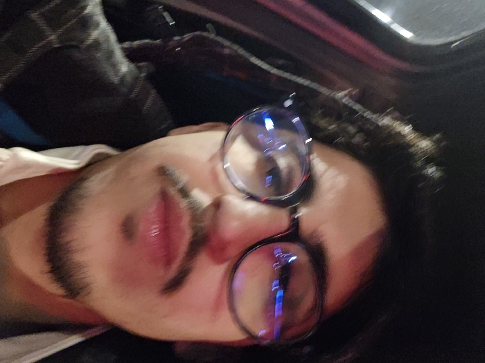 A person with dark hair and a mustache is lying on their side, wearing round glasses. Their face is tilted towards the camera. The background appears dark, possibly inside a car. There is a plaid shirt and a lighter-colored garment visible on the person's shoulder. The light reflects off the lenses of the glasses, creating blue and white flares.