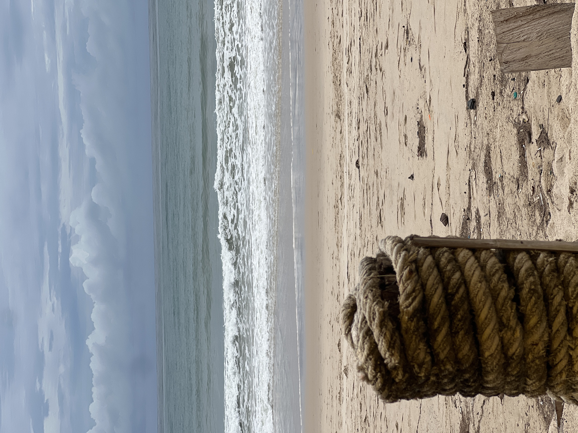 The image shows a coastal scene in an unknown city. The photograph has been rotated 90 degrees counterclockwise. The foreground contains a thick, coiled rope attached to a wooden post. The rope appears worn. Adjacent to the rope and post is a sandy beach strewn with debris. In the middle ground is the surf with foamy waves. In the background, the sky is overcast.
