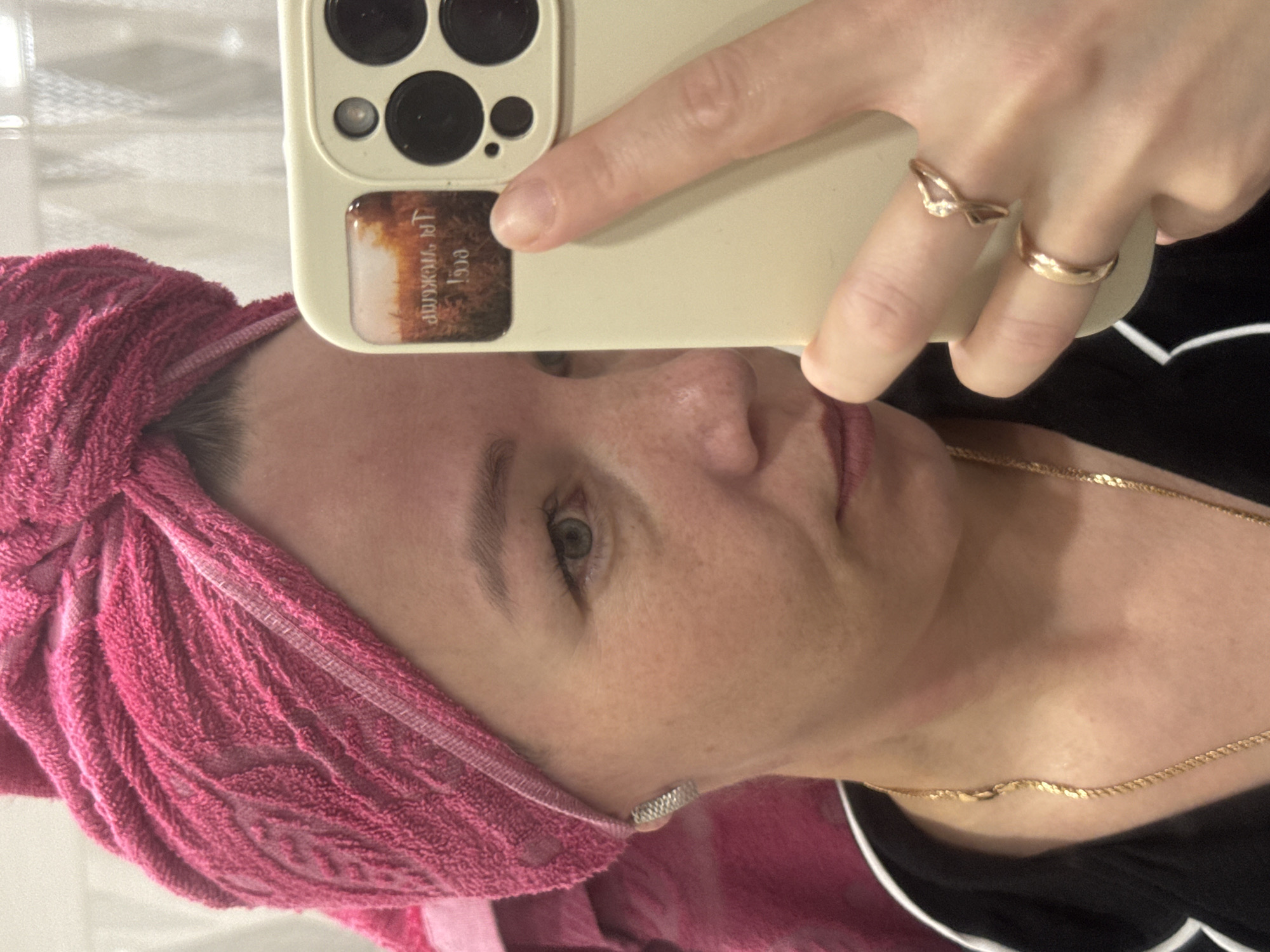 A woman is taking a selfie with an iPhone. She has a pink towel wrapped around her head. She's wearing two rings on the finger of the hand holding the phone and a gold necklace. The iPhone has a sticker on the back that reads "Чайковский, 1936" which is Russian for "Chaikovsky, 1936". Chaikovsky is a city in Perm Krai, Russia.