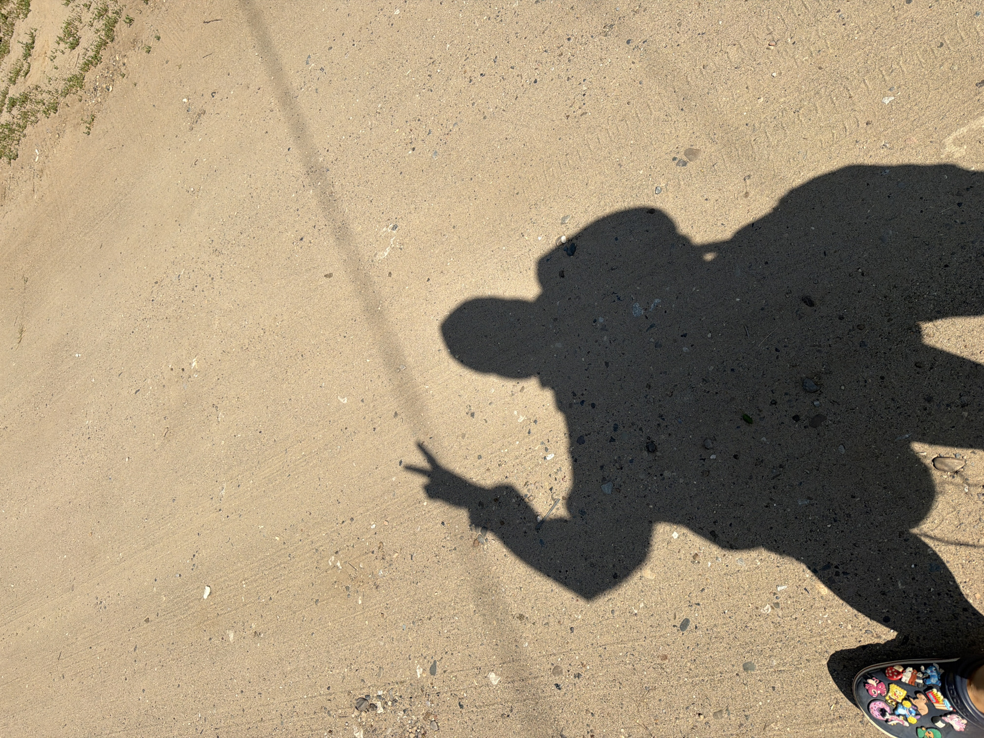 A shadow of a person is cast on a sandy, dusty ground. The shadow shows a person holding up two fingers in a peace sign. The person is wearing shoes decorated with colorful charms. The ground appears to be a path or unpaved road with some small pebbles and sparse dry grass on the right side. A thin, dark line, possibly a wire or thin shadow, runs diagonally across the image from the upper right to the lower left. The lighting suggests it is daytime, with the sun casting a strong shadow. The location is a street in Chaikovsky, Russia.