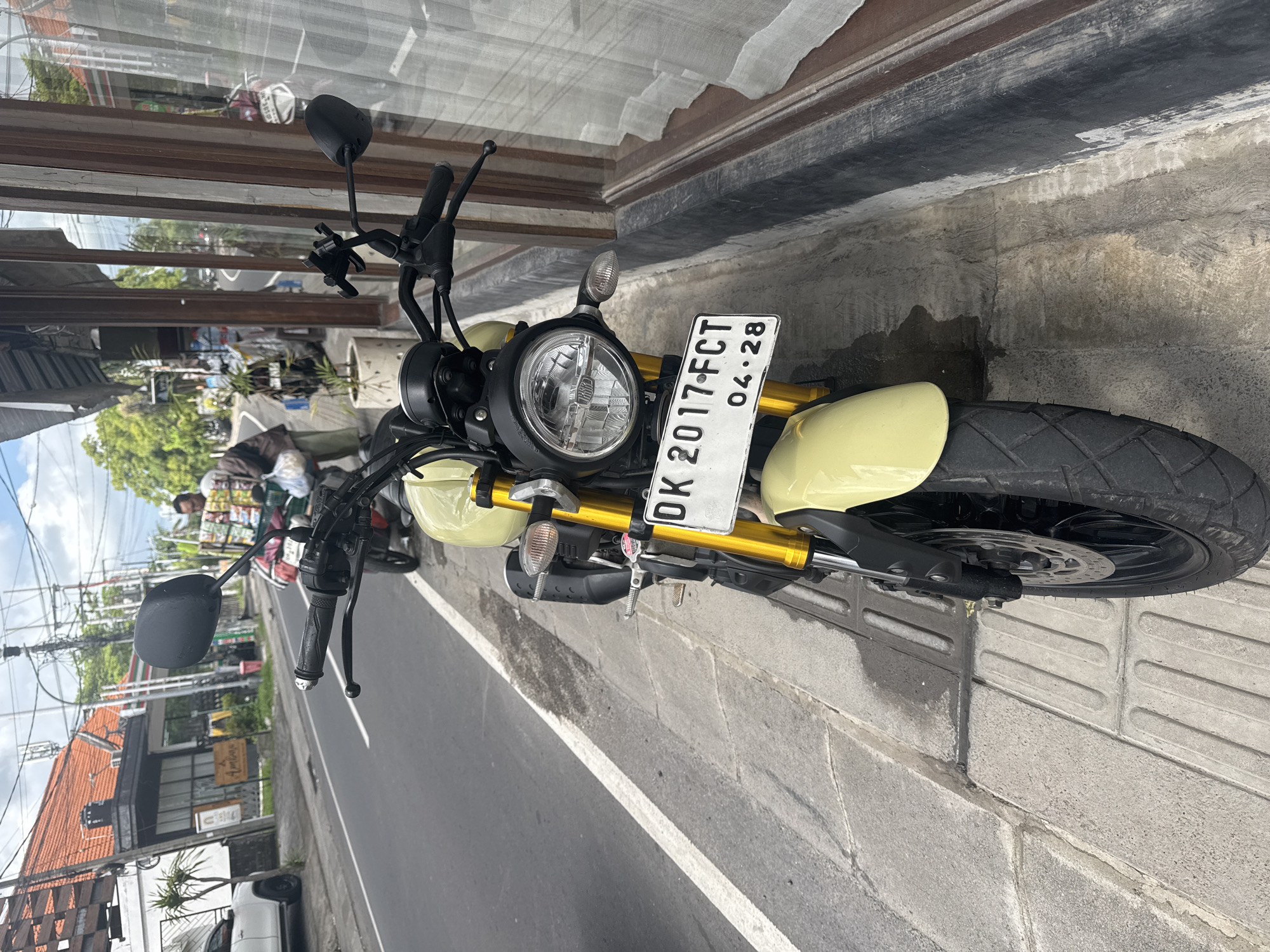 The image depicts a yellow and black motorcycle parked on a sidewalk next to a road in North Kuta, Indonesia. The motorcycle's license plate reads "DK 2017 FCT 04.28." A building with a glass window is to the right of the motorcycle, and its reflection shows a street scene. On the left side of the road, other buildings and pedestrians are visible. The sky is overcast.