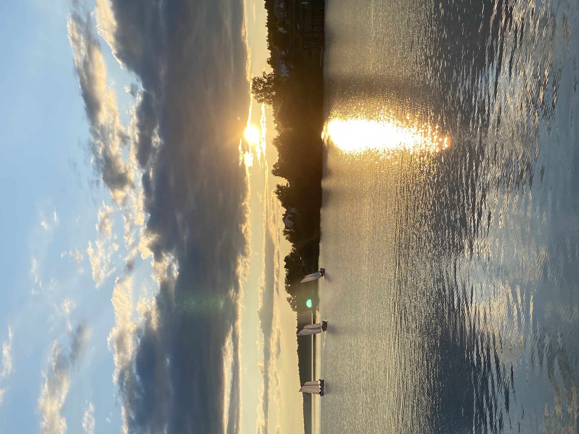 This media file depicts a picturesque sunset or sunrise over a large body of water in Votkinsk, Russia. The sun is low on the horizon, emitting a powerful golden light that creates a shimmering, bright reflection across the water's surface, particularly prominent on the right side of the image. The water itself is calm with gentle ripples, mirroring the colors of the sky and the sun's glare.
The sky above features scattered clouds; some are dark grey, while others catch the golden light, illuminating their edges. Along the distant bank, a line of trees forms a dark silhouette against the bright horizon, with some residential buildings or houses faintly visible behind them.
Three small sailboats, each with white sails, are visible on the water, spaced out across the middle ground. Their sails are clearly defined, suggesting they are either actively sailing or moored. No people are explicitly visible in the boats. The scene evokes a sense of peace and tranquility, characterized by the warm, golden hues of the sun. There is no visible text.