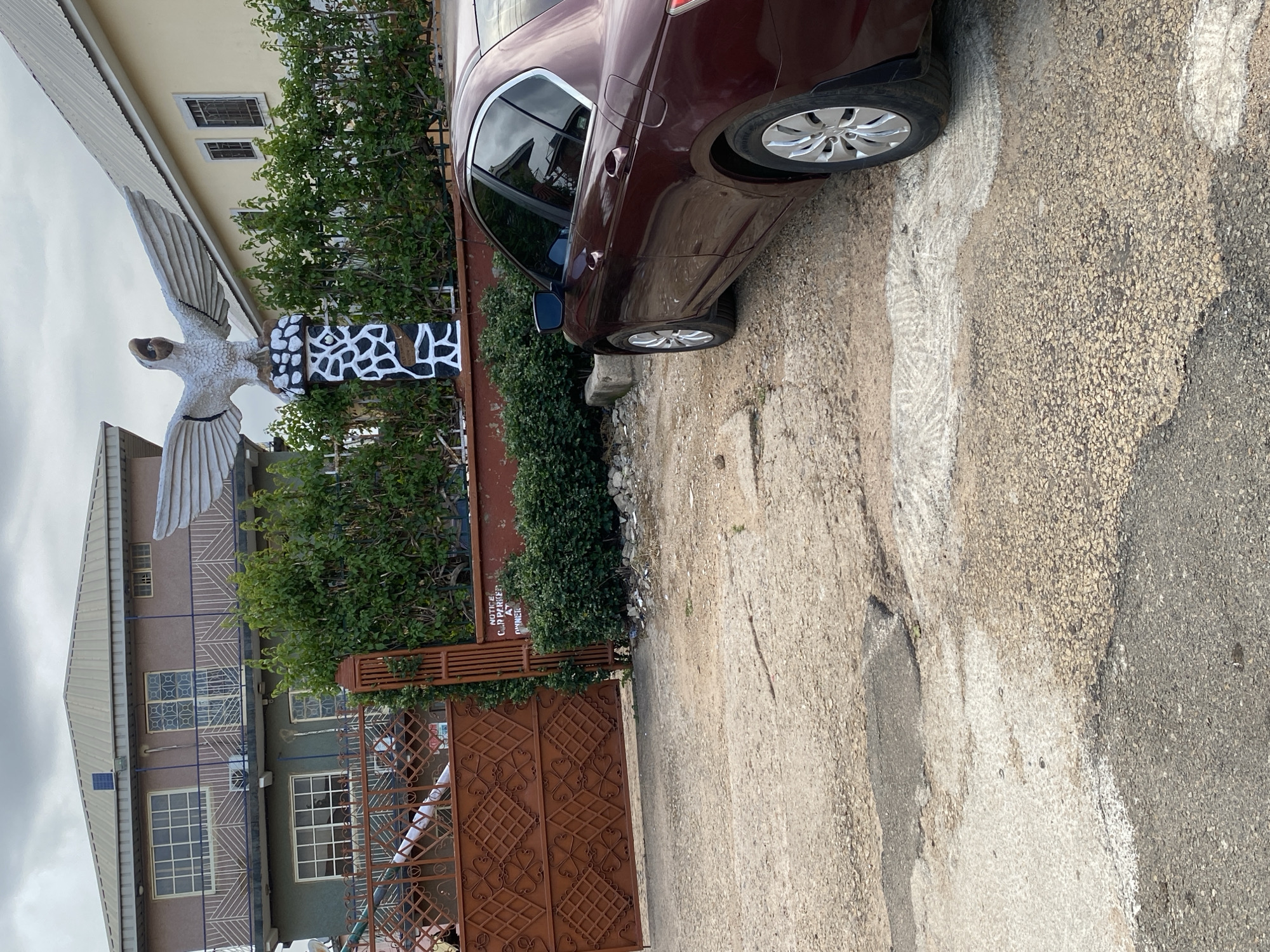 A maroon car is parked on a gravel and concrete surface. The car is angled, with its rear facing the camera. A white sculpture of a bird with its wings spread is visible to the left of the car, in front of a building. The building appears to be a multi-story structure with windows and a patterned facade. There is also a brown metal gate and lush green foliage in the scene. The ground is rough and uneven, with some white markings, possibly indicating parking spaces. The sky is overcast, suggesting a cloudy day. The image appears to be taken outdoors in a residential or commercial area. There is a sign visible on the gate that says "NOTICE CAR PARKED AT OWNER'S RISK". The overall scene is static, with no apparent activity or event taking place. The lighting suggests it is daytime.