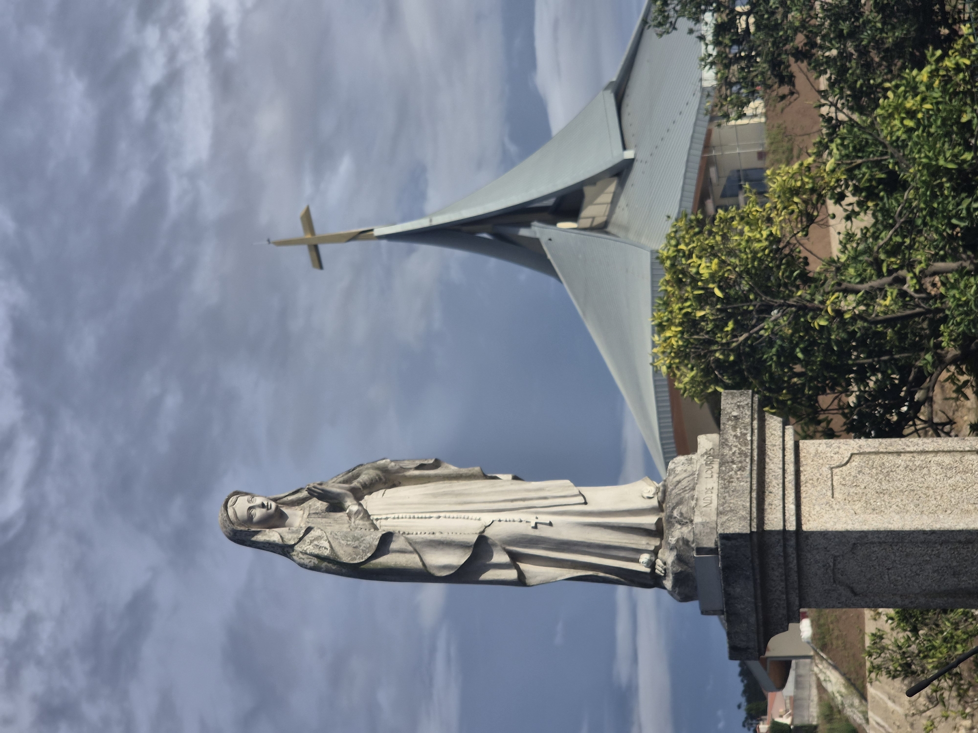 The image shows a statue of the Virgin Mary in front of a modern church with a distinctive roofline and a golden cross. The statue is made of stone and depicts the Virgin Mary with her hands clasped in prayer. She is wearing a draped robe and a veil, and a rosary hangs from her neck. The statue stands on a stone pedestal with an inscription that reads "ND DE LOURDES." In the background, the church features a large, metallic roof with sharp angles, and a golden cross is prominently displayed at its apex. Lush green trees are visible in the foreground, partially obscuring the lower part of the church. The sky is filled with dramatic, cloudy formations. The inscription "ND DE LOURDES" on the pedestal likely refers to Our Lady of Lourdes, and the statue's presence suggests a Catholic religious site. This image was captured in Póvoa de Varzim, Portugal, specifically in front of the Santuário de Nossa Senhora da Apresentação (Shrine of Our Lady of Presentation).