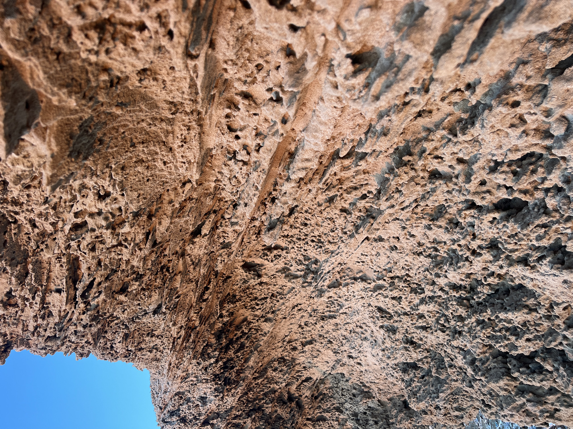 The media file is a close-up, low-angle shot looking up at the underside of a rocky overhang or archway. The rock surface is rough, porous, and textured, with many small holes and crevices, suggesting it might be weathered volcanic rock or a similar geological formation. The color of the rock is predominantly a warm, earthy brown with lighter tan and darker grey-brown shadows, indicating it's likely illuminated by natural light. The lighting suggests it is daytime, possibly late afternoon given the warm tones and shadows.
A portion of a clear, bright blue sky is visible through the opening of the arch, providing a stark contrast to the textured rock. The image is a detail shot, focusing entirely on the geological formation and the sky. There are no people, man-made objects, or specific activity depicted. The setting appears to be a natural outdoor environment, possibly a coastal area or a cave entrance, given the weathered rock and open sky. The lack of any recognizable landmarks or human presence implies a focus on the natural textures and forms of the landscape. No text is visible in the media file.