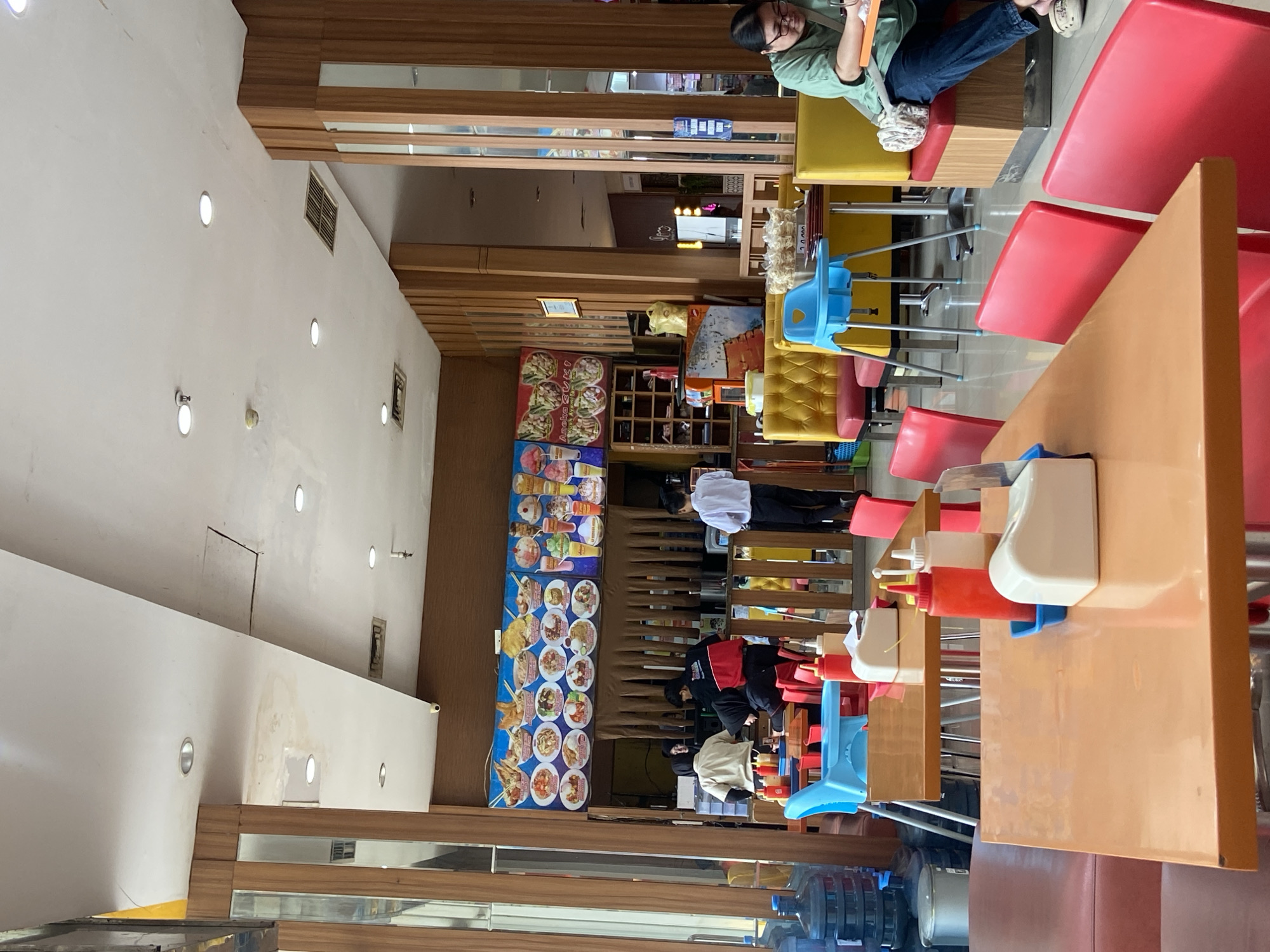 The image depicts the interior of a casual dining establishment in Kudus, Indonesia, likely a fast-food restaurant or a food court stall. The scene is brightly lit and features a variety of seating arrangements, including orange tables with red chairs, and yellow booth seating. A large menu board displaying various food items and drinks is prominently displayed on the back wall, above a counter area where employees are visible. Several employees, dressed in uniforms or casual attire, are attending to customers or preparing food behind the counter. In the foreground, tables are set with condiments like ketchup and chili sauce dispensers, and tissue holders. Water cooler bottles are visible on the left side of the image, suggesting a self-service water station. The overall ambiance is one of a busy, everyday eatery.