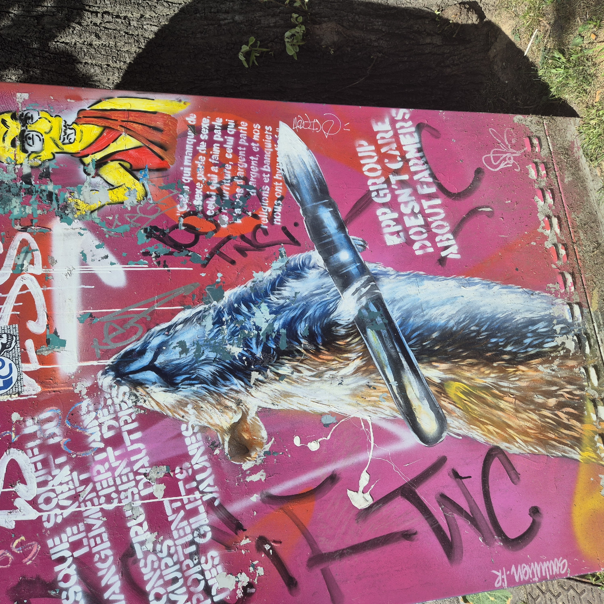 The image shows a rectangular concrete structure covered in graffiti in Strasbourg, France. The graffiti includes a cartoon figure, a blue wolf-like creature with a bullet or pen drawn across it, and text in French and English. The French text mentions “sex, food, money, religion, and bankers” and the English text says “EPP GROUP DOESN’T CARE ABOUT FARMERS”. There is also the word “TWC” spray-painted in large letters. Some of the paint is peeling off, and the structure is located near a tree or bush, with foliage visible in the background. The ground appears to be concrete or stone.