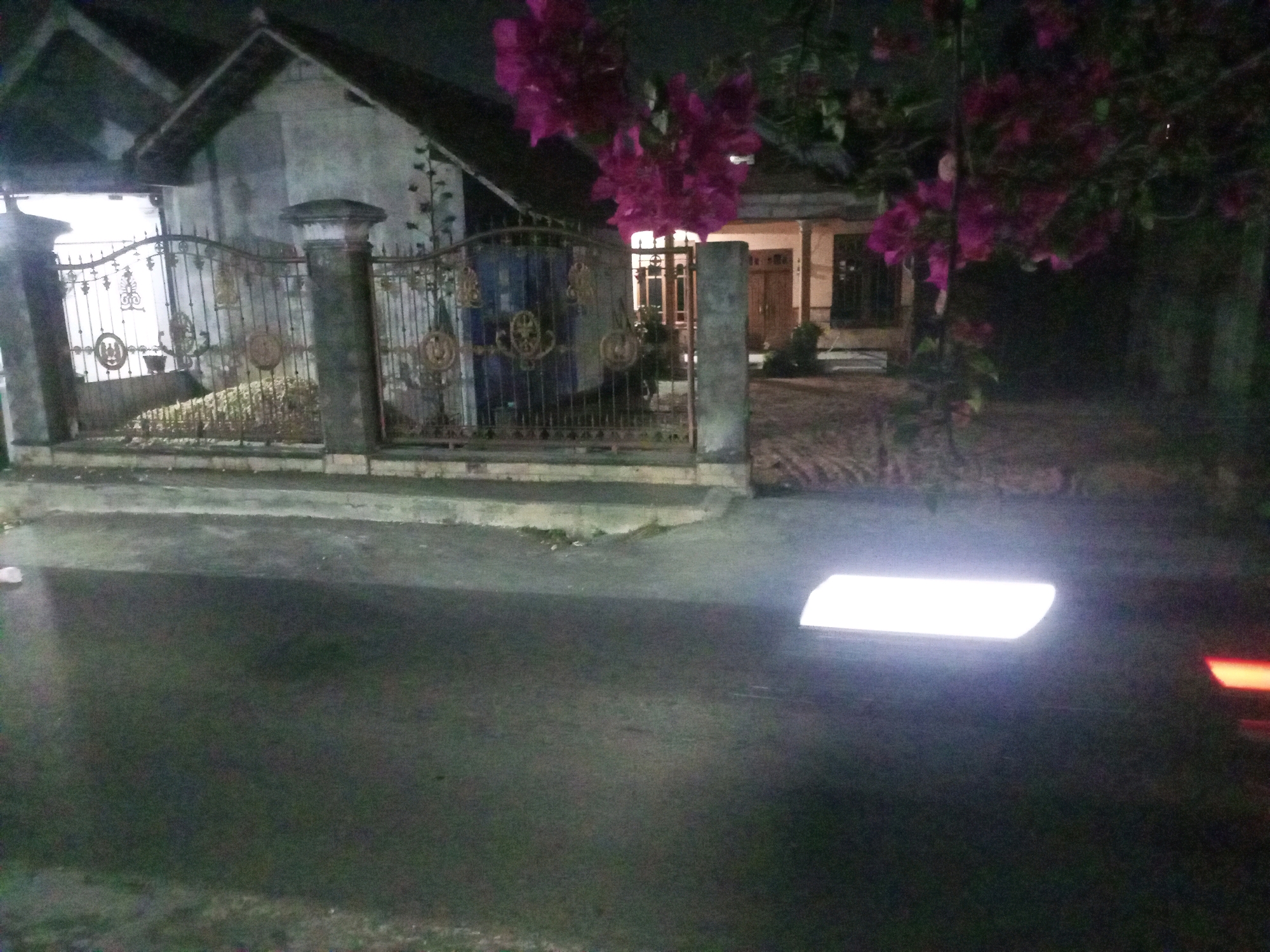 A night-time exterior scene in Ngasem, Indonesia, features a residential property with a decorative metal gate in the foreground, partially obscuring the house and surrounding vegetation. The house is a single-story structure with a pitched roof and a warm-toned facade, featuring wooden doors and windows. Vibrant pink bougainvillea flowers cascade from a tree, adding a splash of color to the dark surroundings. The ground in front of the gate appears to be paved, transitioning to a darker, possibly asphalt, surface in the foreground. A bright, rectangular light source is visible on the darker surface in the lower right quadrant of the image, suggesting a vehicle or external lighting. The overall impression is one of a quiet, domestic setting illuminated by artificial light against the backdrop of the night.