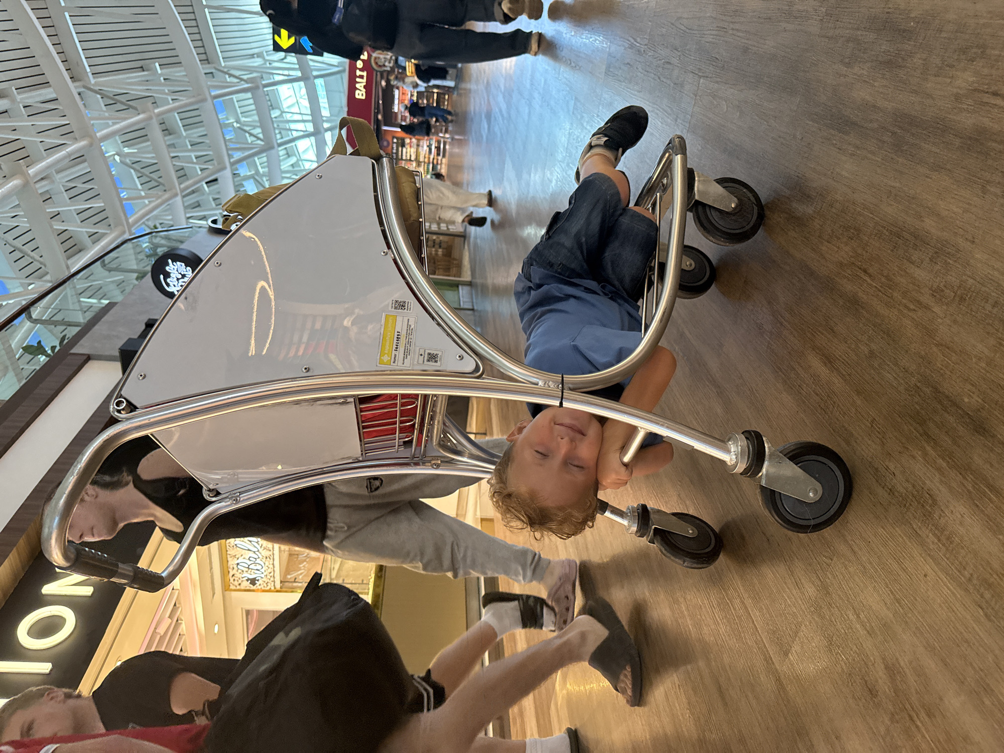 A young Caucasian boy with blonde, curly hair and a blue shirt is lying on his stomach inside a metal luggage cart. His head is resting on the cart's handle, and his eyes are closed, appearing relaxed or asleep. He is wearing denim shorts. The cart has silver metal tubing and black wheels. The scene takes place on a polished wooden floor in what appears to be an airport terminal, with other people and shops in the background. The overall lighting suggests an indoor environment, possibly Kuta, Indonesia, given the "BALI" signage visible in the background. The boy's playful pose suggests a moment of rest or amusement during travel.