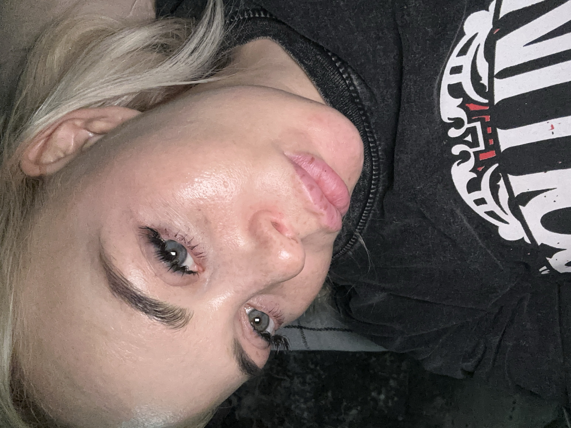 A close-up, angled portrait captures a person with light blonde hair, positioned as if lying down and looking slightly upwards. The individual has light blue-green eyes enhanced by long, dark, voluminous eyelashes, likely extensions, and neatly groomed, dark eyebrows. Their skin appears clear with a natural, subtle sheen across the forehead and cheek. The lips are full and slightly parted, showing some natural texture.
Blonde hair cascades around the head, partially obscuring an ear on the left side of the frame. The person is dressed in a dark grey or black hooded top, possibly a sweatshirt or jacket, with a visible zipper detail near the neckline. A prominent graphic design on the fabric of the garment features an intricate white pattern with subtle red accents, hinting at a stylized emblem or artistic motif, though no specific text is legible.
The setting appears to be indoors, indicated by the diffused lighting and the dark, out-of-focus background that offers no specific location cues. The overall impression is a focused, almost intimate shot, likely a selfie or a close-up photo, with the subject exhibiting a calm and neutral expression.