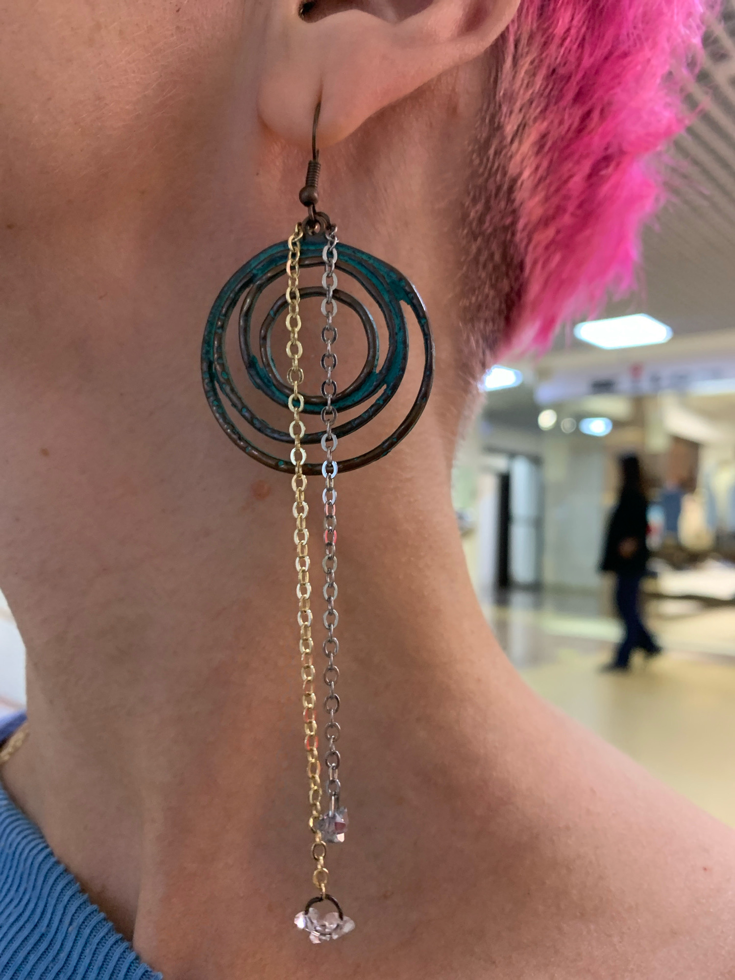The image is a close-up, shoulder-level shot of a person's left ear and neck. The person has bright pink hair. They are wearing a long, multi-layered hoop earring with chains of alternating gold and silver tones, adorned with small clear crystals at the bottom. The earring is attached to a dark-colored ear hook. The person is wearing a blue, ribbed garment. The background is blurred but appears to be an indoor public space with fluorescent lighting, possibly a mall or hospital corridor, with a blurred figure of another person walking in the distance. The image does not contain any information about the city of Arkhangelsk, Russia.