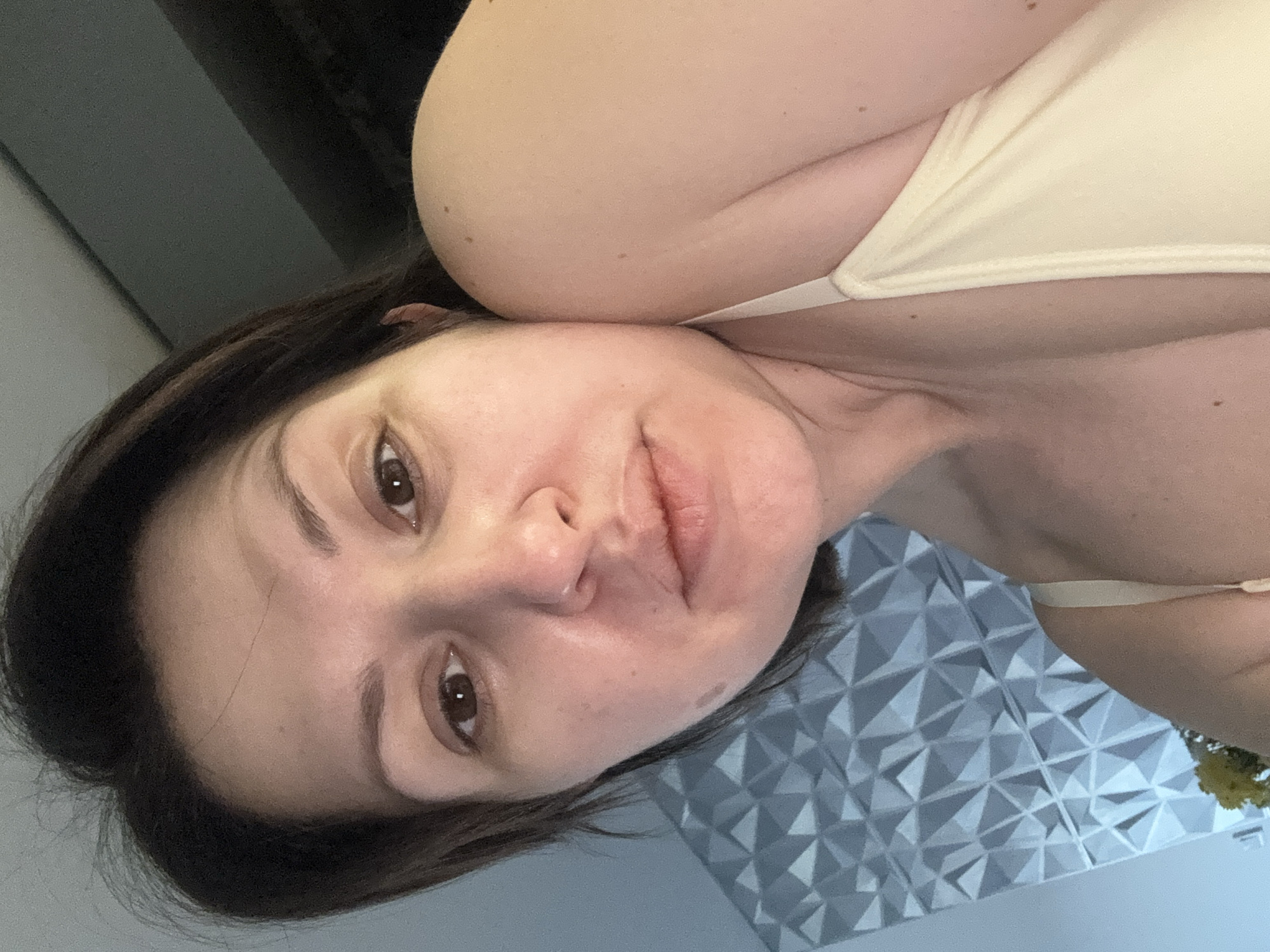 The image is a close-up selfie of a woman with brown hair and brown eyes. She is fair-skinned and appears to be wearing a light-colored top with straps. The background includes a textured, geometric wall pattern in shades of gray and white. The location is not identifiable as Sofia, Bulgaria, without further visual cues or information.