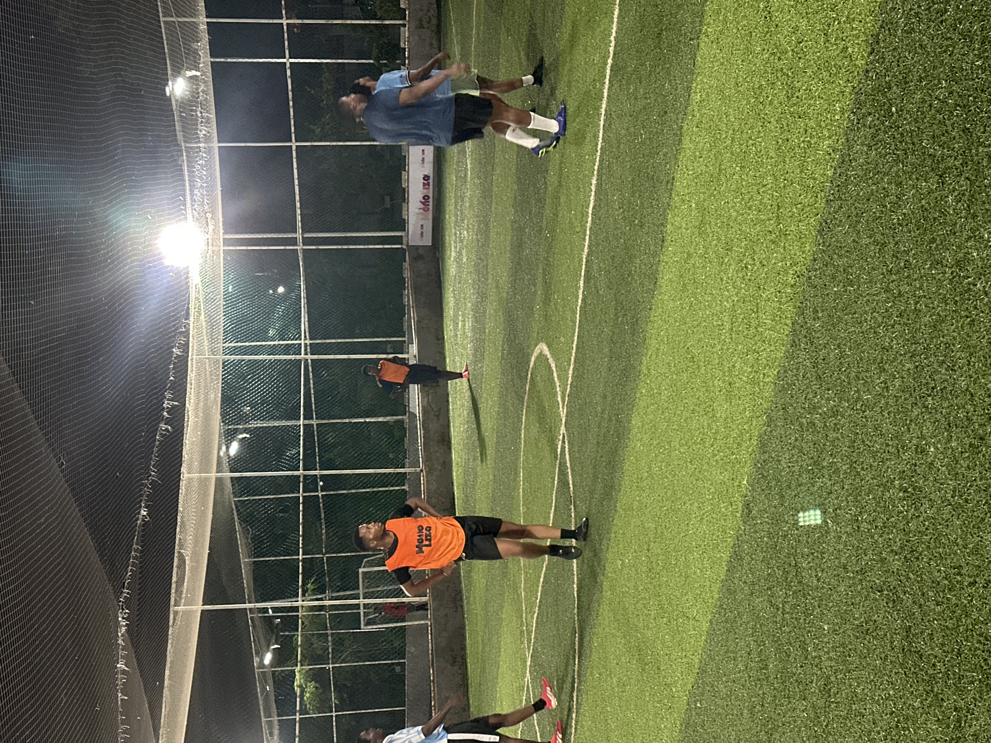 The image depicts an outdoor artificial turf football field at night in Abuja, Nigeria. The field is illuminated by overhead floodlights, creating bright spots and shadows. Several individuals, appearing to be young men, are present on the field, some in athletic wear and some wearing orange bibs. One individual in the foreground is wearing an orange bib with the inscription "Metro Liga" and black shorts. Another individual to his left is wearing a light blue and white striped jersey with black shorts. Further in the background, other individuals are visible, some in orange bibs. The field is bordered by a fence and netting, suggesting it is an enclosed sports facility. The overall atmosphere suggests an informal football game or practice session is underway.